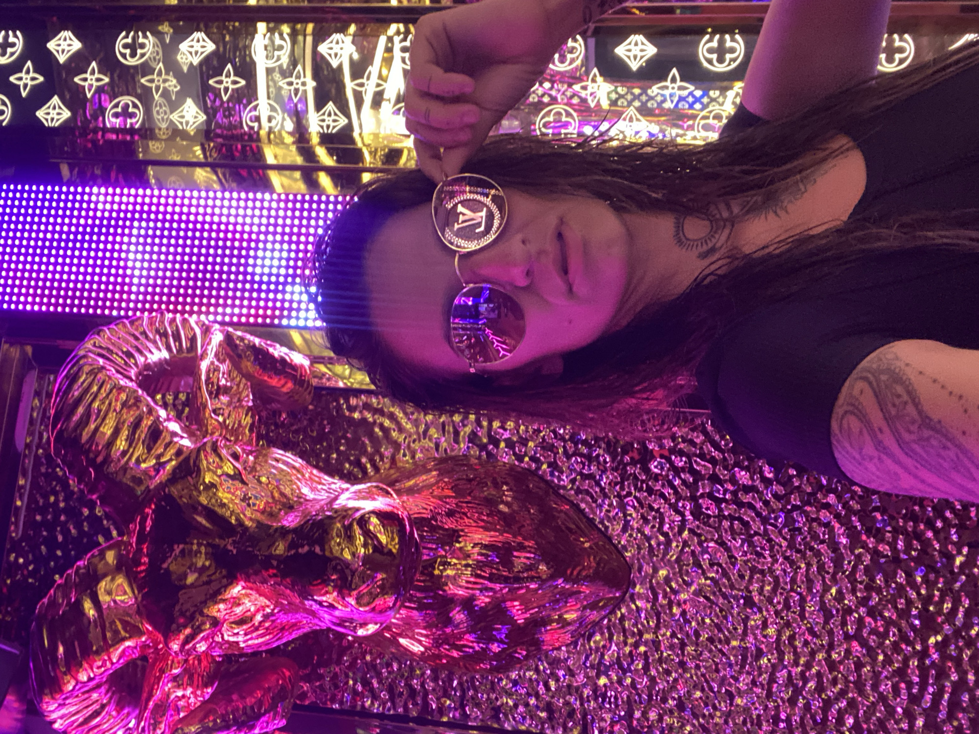 The image shows a woman wearing round sunglasses with the Louis Vuitton logo on the frame, posing in what appears to be a luxurious interior in Đồng Nai, Vietnam. The backdrop is adorned with a gold and purple neon lighting scheme, featuring Louis Vuitton's iconic quatrefoil patterns. A large, golden, abstract sculpture is present on the left side of the image. The woman has dark hair and several tattoos visible on her arm and neck. The texture of the wall is mirrored and distorted due to the lighting.