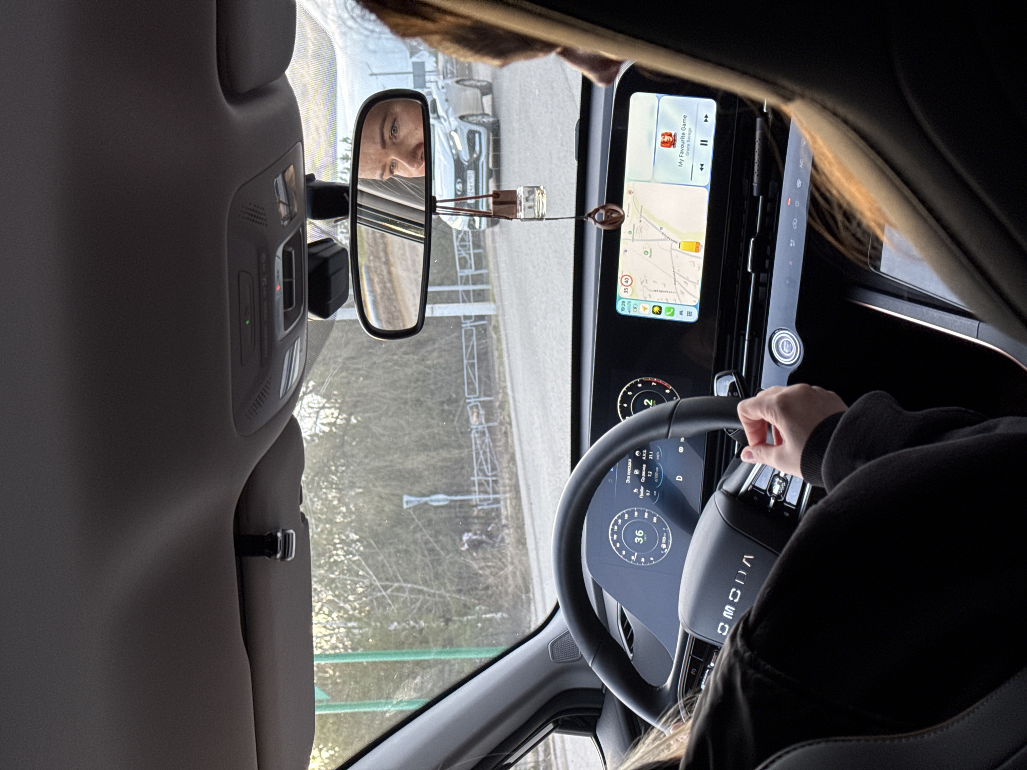 The image shows the interior of a car, with the driver visible in the rearview mirror. The infotainment screen displays a map and music information. The car's dashboard and steering wheel are also visible, with the speedometer reading 36 km/h. The navigation information shows some Russian characters and numbers 35 and 40, possibly a speed limit. The environment outside the car consists of trees and possibly a building in the distance. The image includes what seems to be a small perfume bottle hanging from the rearview mirror.
