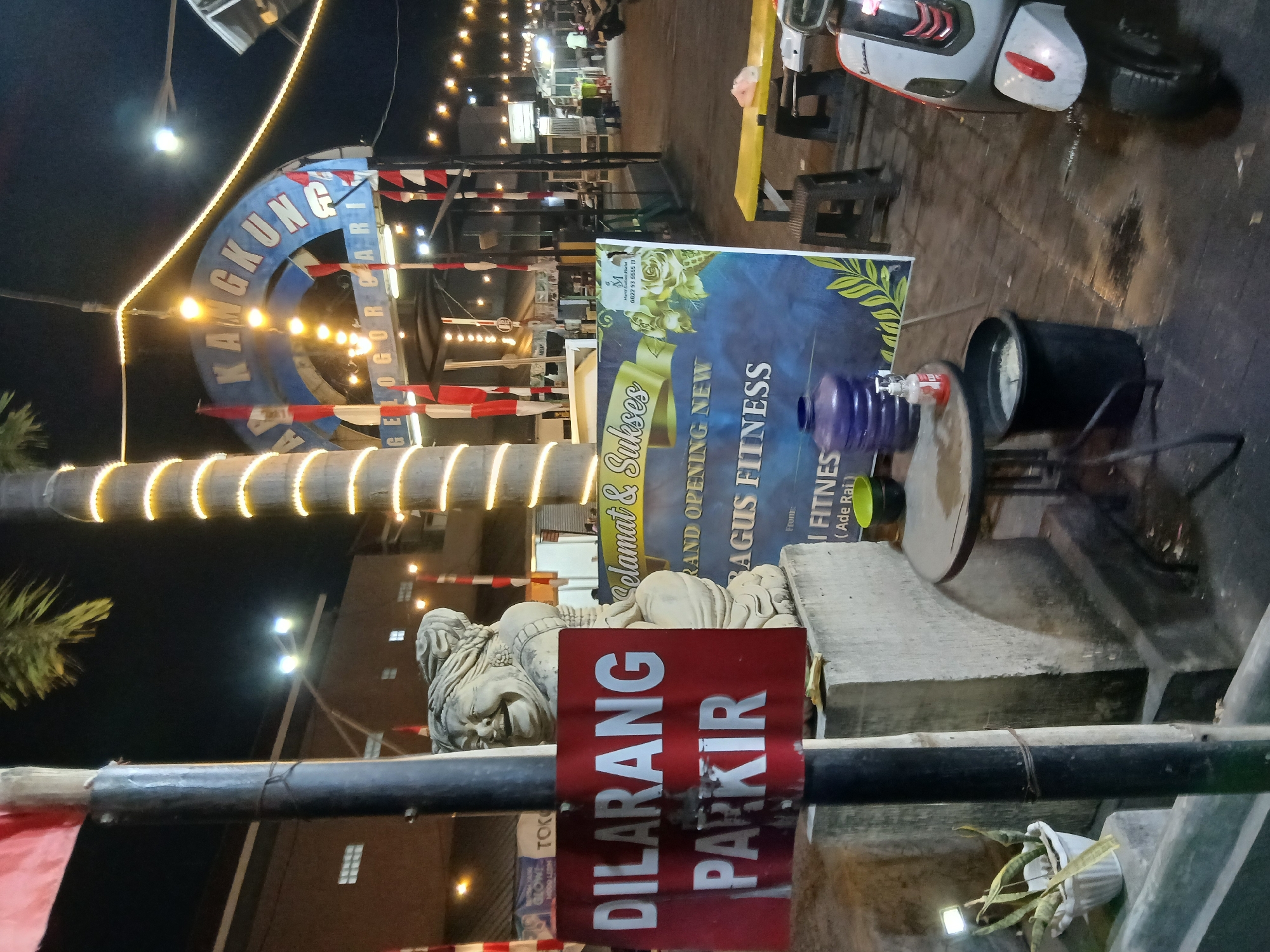 In Denpasar, Indonesia, at night, a statue of a laughing woman stands next to a pole with a red sign that reads "DILARANG PARKIR" (No Parking). A large banner, advertising "SELAMAT & SUKSES GRAND OPENING NEW SAGUS FITNESS," is displayed behind the statue, featuring floral imagery. A water cooler jug and a small round table with a black bucket are positioned nearby. The scene is illuminated by strings of lights and streetlights, casting a warm glow on the wet pavement. A scooter is parked to the right of the frame. In the background, shops and other establishments are visible, some adorned with red and white flags.