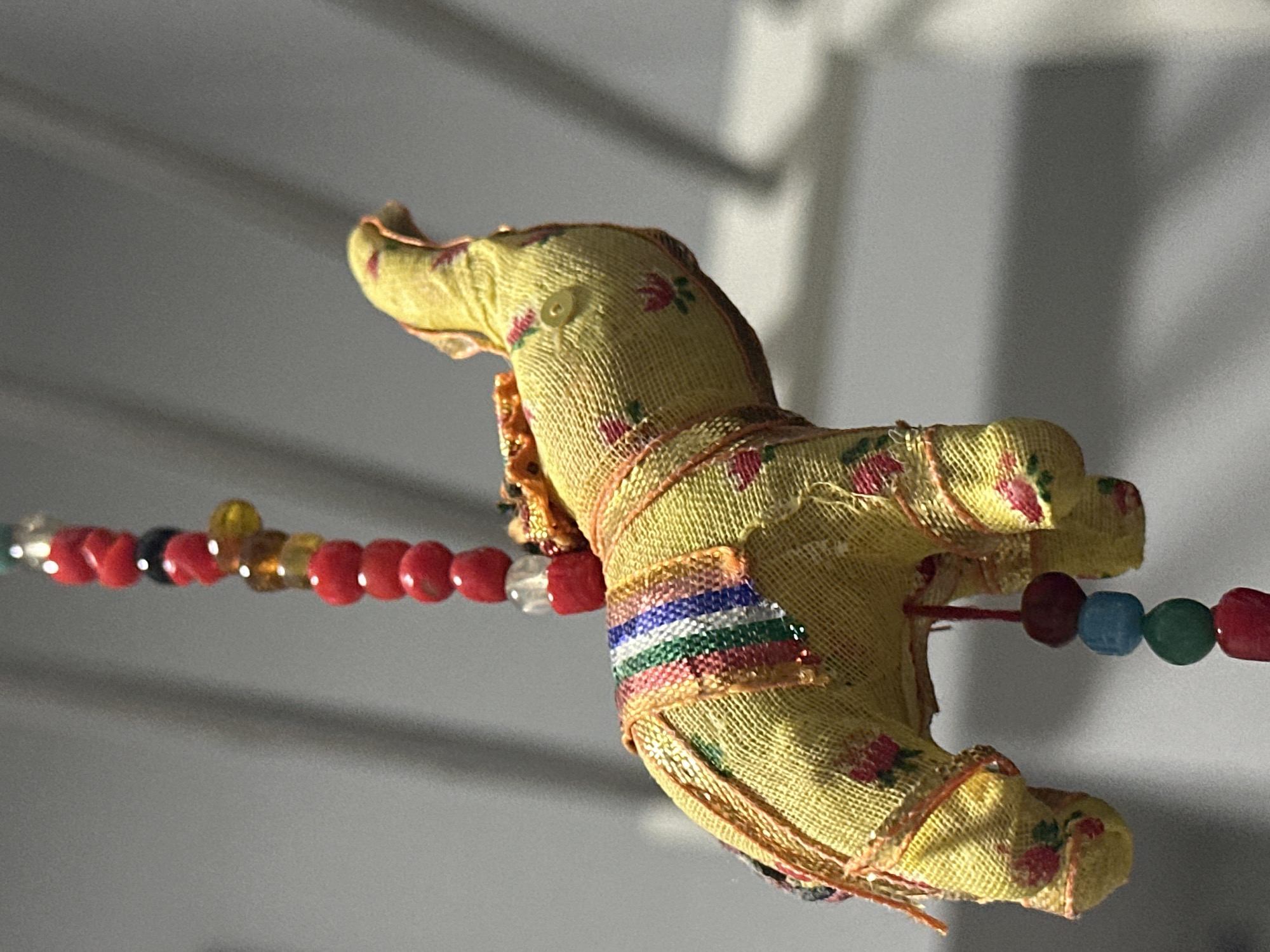 A yellow fabric elephant, adorned with a beaded necklace and dangling beads, hangs suspended against a grey, out-of-focus background. The elephant is decorated with a striped sash and floral patterns. The necklace is composed of red, black, and amber beads, while the dangling beads are red, blue, green, and red. The scene is likely indoors, given the lack of natural light and the presence of what appears to be a ceiling or wall with a striped shadow. The image does not provide any specific clues about its location in the city of Nevinnomyssk, Russia.