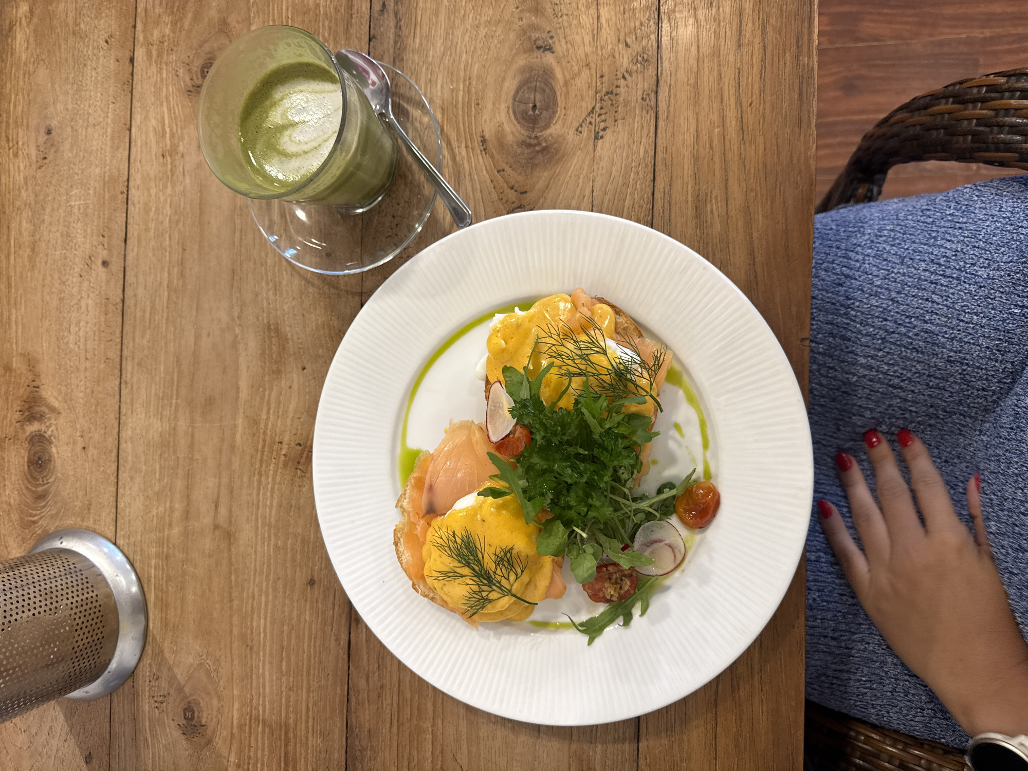 The image shows a top-down view of a meal and drink served on a wooden table. The main subject is a white plate with what appears to be Eggs Benedict, topped with salmon and a small salad of greens and cherry tomatoes. A green sauce accents the edge of the plate. Next to the plate is a clear glass of a green beverage, possibly a matcha latte, with a small silver spoon resting on its saucer. To the right of the plate, a person's hand with red nail polish rests on a blue textile covering what looks like a chair made of woven material. A smartwatch is visible on the person's wrist. A metal object is on the bottom left of the table. The background suggests the location might be in Ubud District, Indonesia, due to the style of the food and the presence of a matcha latte, which is a popular beverage in the area.