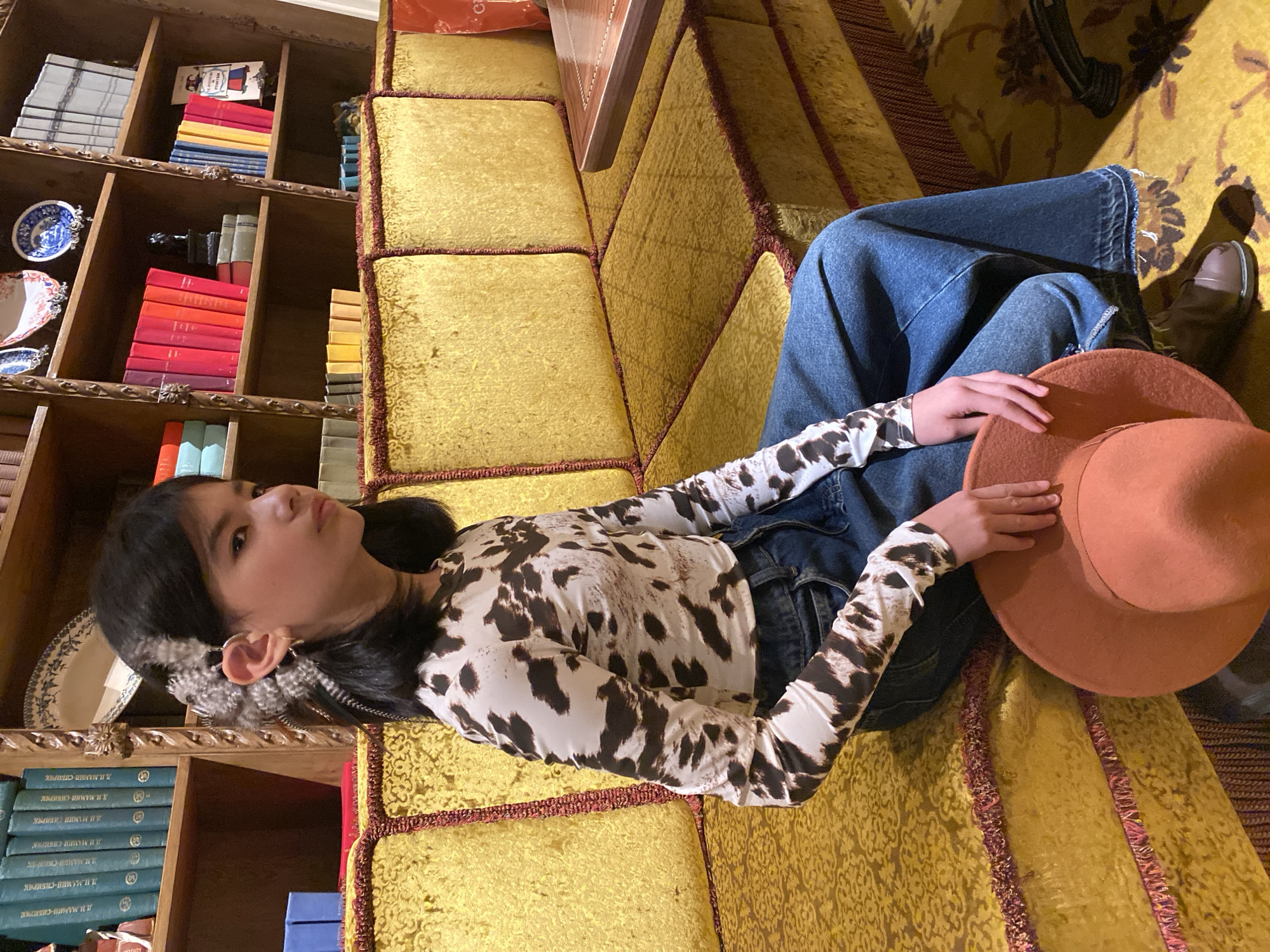 A young person, likely a girl or young woman, is pictured lying on a plush, gold-colored upholstered surface, possibly a bench or floor cushions, with red trim. She wears a long-sleeved top with a black and white cow print, loose blue jeans, and holds a rust-orange, wide-brimmed hat. Her dark hair is styled around her face, and she has unique feather-like adornments near her left ear. Her gaze is directed upwards and slightly to the left with a neutral, contemplative expression.
The setting appears to be an indoor space with a traditional aesthetic, possibly a library or study. A large, ornate wooden bookcase lines the wall behind her, filled with numerous books. Many books have red or orange spines, while others are blue or light-colored, suggesting a collection of classic literature or a well-curated library. Decorative blue and white ceramic plates or dishes are interspersed among the books on the shelves. Partial views of other wooden furniture, possibly a table, are visible in the upper right.
Several books display Cyrillic text, specifically identifying the author "Д.И. МАМИН-СИБИРЯК" (D.I. Mamin-Sibiryak), a renowned Russian writer. This detail, combined with the provided context, places the scene in Russia, potentially Yekaterinburg, the author's birthplace. The lighting is indirect, indicating an indoor shot without strong natural light, or possibly an evening setting. The overall mood is calm and introspective.