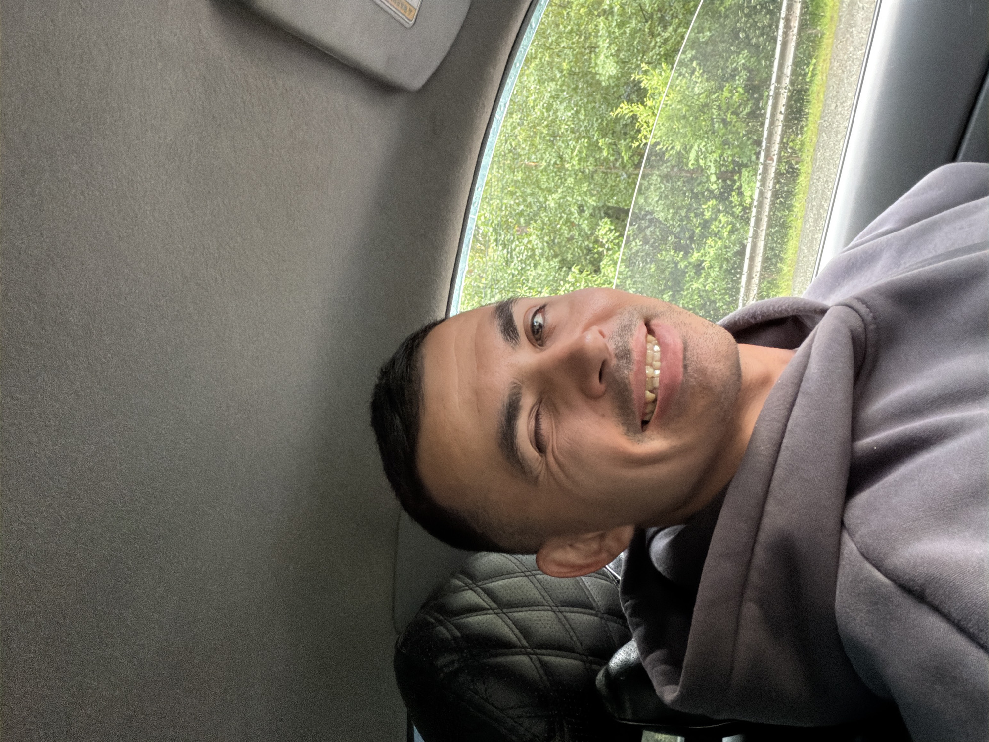 The image shows a close-up selfie of a man in a car. The man is smiling and looking towards the camera. He is wearing a gray hooded sweatshirt. His left eye is partially closed, giving him a playful expression. In the background, through the car window, lush green trees and a rainy day can be seen. The interior of the car is visible, with a gray headliner and a black, quilted leather car seat. The scene suggests a casual, possibly spontaneous, moment captured while traveling. The man's expression and the soft lighting indicate a relaxed or happy mood. There is no visible text in the image. The location context provided is Izhevsk, Russia.