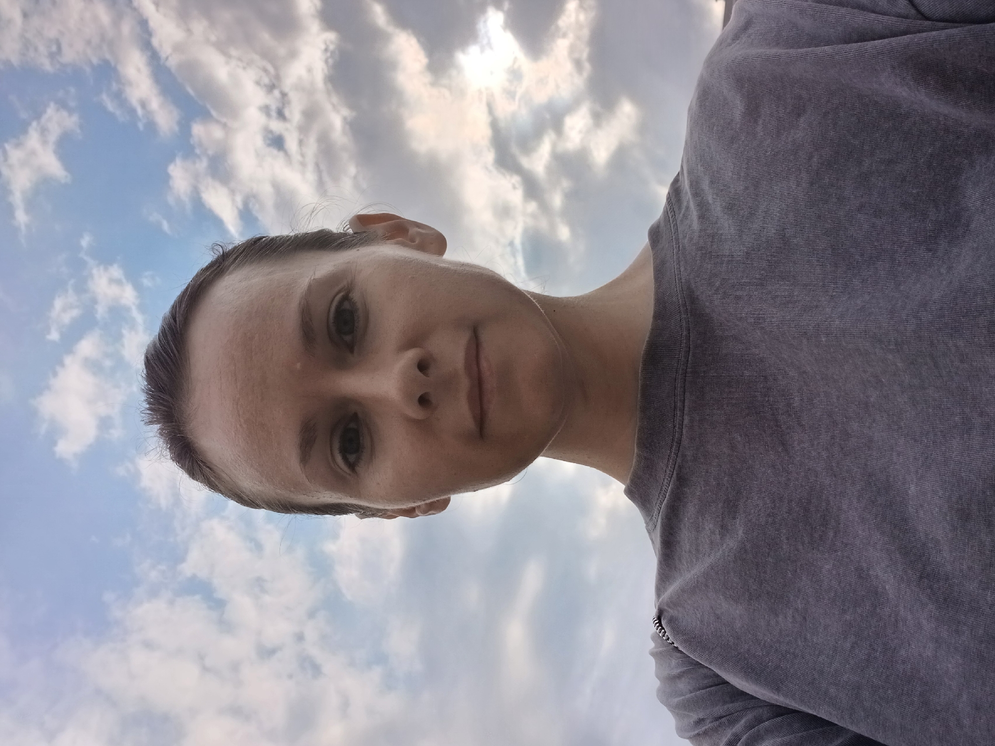 This is a close-up, low-angle selfie of a woman with her hair pulled back, facing the camera. She is wearing a grey sweatshirt with a zipper detail on the left shoulder. The background consists of a cloudy sky. The image is taken outdoors, potentially in Yekaterinburg, Russia, given the context.