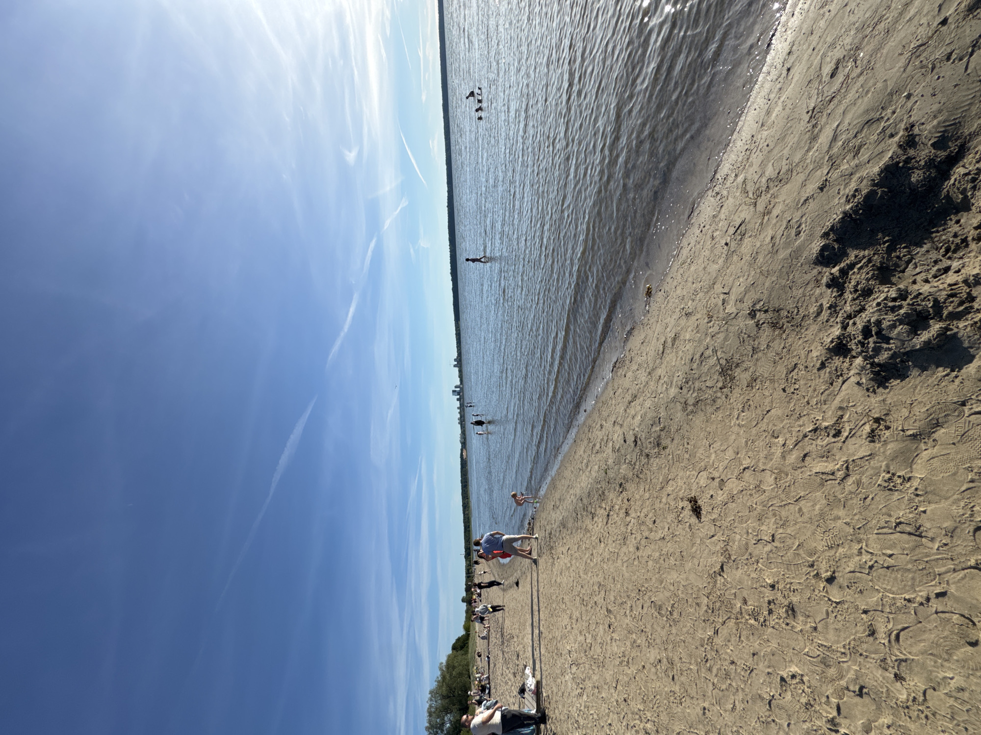 This media file depicts a sunny day at a beach in Tallinn, Estonia. The scene is oriented vertically, with the sky occupying the left side and the beach and water on the right. A wide expanse of water, likely the Baltic Sea, stretches across the upper portion of the image, with gentle waves rippling its surface under the bright sunlight. The sky is a clear blue with some wispy white contrails.
The foreground is a sandy beach with footprints clearly visible, indicating recent activity. Several people are scattered across the beach and in the shallow water. On the beach, a man in a blue shirt and grey shorts is standing near a woman in red, possibly interacting or walking together. Further back, other individuals are seen enjoying the beach, some walking and others appearing to relax.
In the water, a few people are visible. One person is wading in the shallow area, while further out, a group of what appear to be birds or people are spread out on the water. The shoreline is defined by the wet sand where the water meets the beach. In the distance, along the horizon, a line of trees and buildings suggest the presence of the city or a park area. The overall impression is one of a pleasant, leisurely day outdoors, likely in the late morning or early afternoon given the bright sunlight and shadows. There is no visible text in the image.