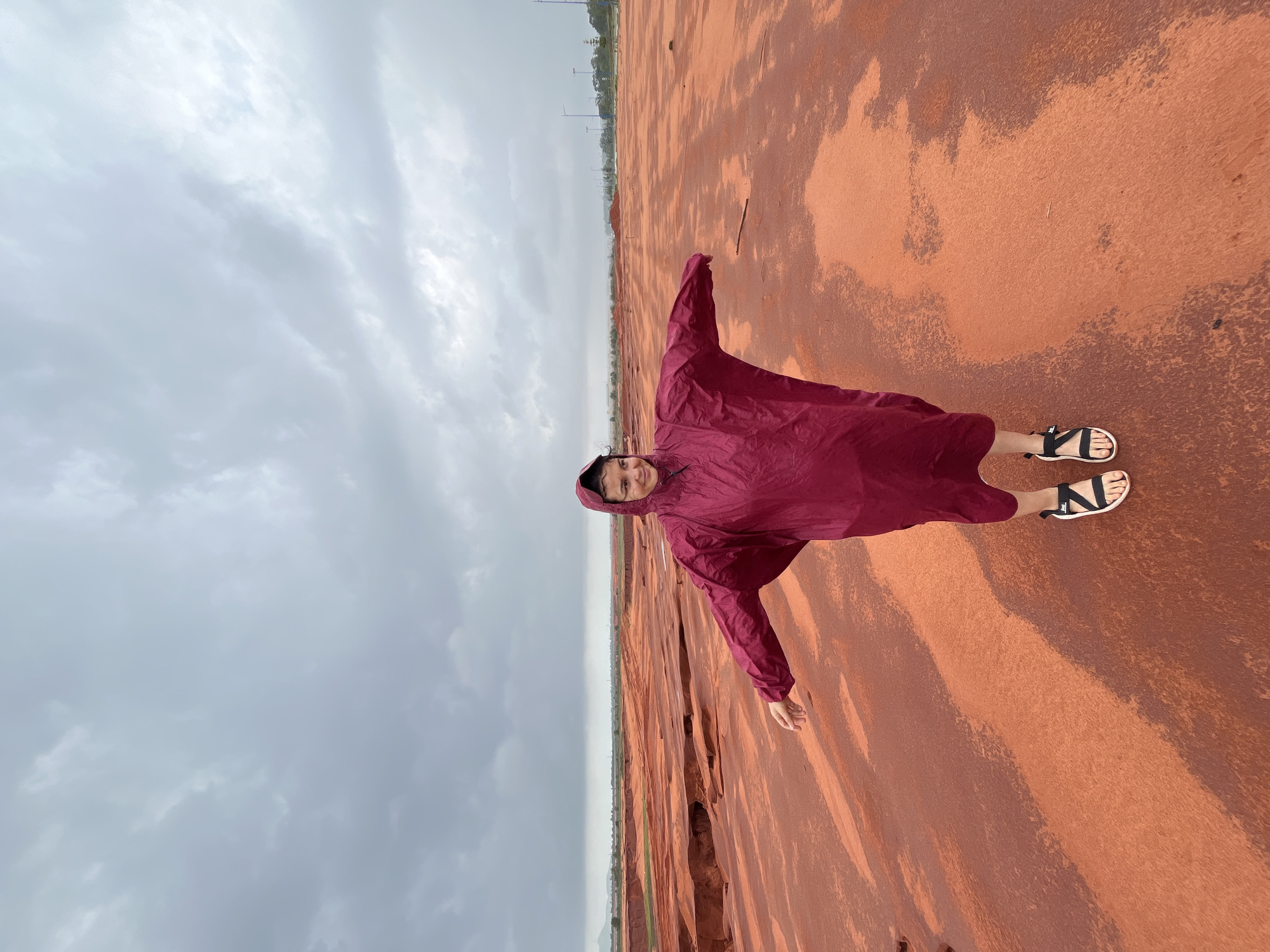 A person stands in a vast, open area of red clay under a cloudy sky. The person is wearing a maroon poncho and black sandals, with their arms outstretched to the sides. The ground is a deep red, with patches of lighter color and some evidence of moisture. In the distance, there is a horizon line with some greenery and structures, suggesting an urban or developed area. The overall impression is of a natural landscape under an overcast sky, possibly indicating rain or recent rainfall. The location is identified as Mui Ne, Vietnam, which is known for its distinctive red sand dunes.