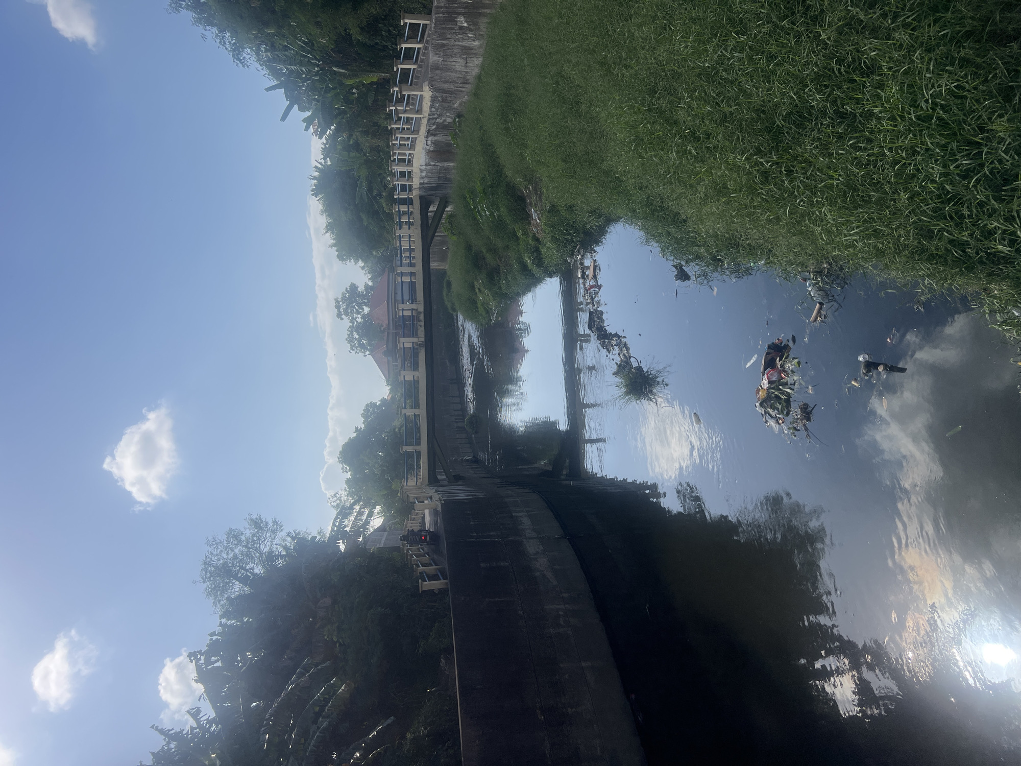 An outdoor, slightly elevated, eye-level, long shot captures a sunny day over a canal in Denpasar, Indonesia. The canal runs through a built-up area, with concrete walls lining its sides. The water is dark and reflects the sky and surrounding foliage. Debris, including plastic bags and bottles, floats on the surface of the water. Dense green grass grows along the right bank of the canal. A bridge spans the canal in the middle ground, with a yellowish railing and concrete supports. A motorcycle carrying one rider is visible on the bridge, heading away from the camera. Buildings with red-tiled roofs are visible behind the bridge and trees on the left side of the image. The sky is bright blue with a few fluffy white clouds. The overall scene depicts a canal in an urban or semi-urban environment with signs of pollution.