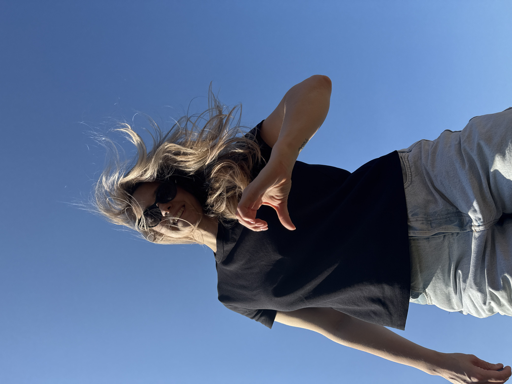 A young woman with blonde hair and sunglasses is photographed from a low angle against a clear blue sky. She wears a black t-shirt and light-wash denim jeans. Her hair is slightly windblown. One of her hands is raised, forming a heart shape with her fingers. The location is specified as Chaikovsky, Russia, though this is not discernible from the image itself.