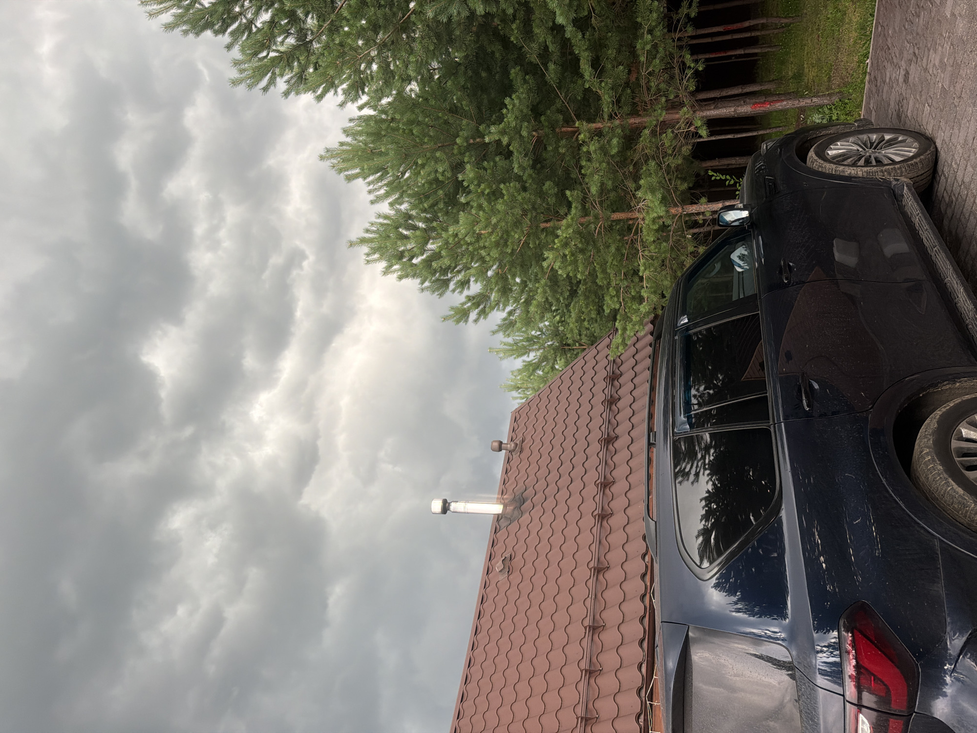 The image captures an outdoor scene on a cloudy day in the village of Kolyutkino, Russia. A dark-colored SUV is parked on a brick-paved surface. To the left of the SUV, a portion of a building with a brown, tiled roof and a stainless steel chimney is visible. Behind the building and the car, a dense line of pine trees stands. The sky is overcast with heavy, gray clouds, suggesting an impending storm or a gloomy day. The overall impression is one of a rural or suburban setting under challenging weather conditions.