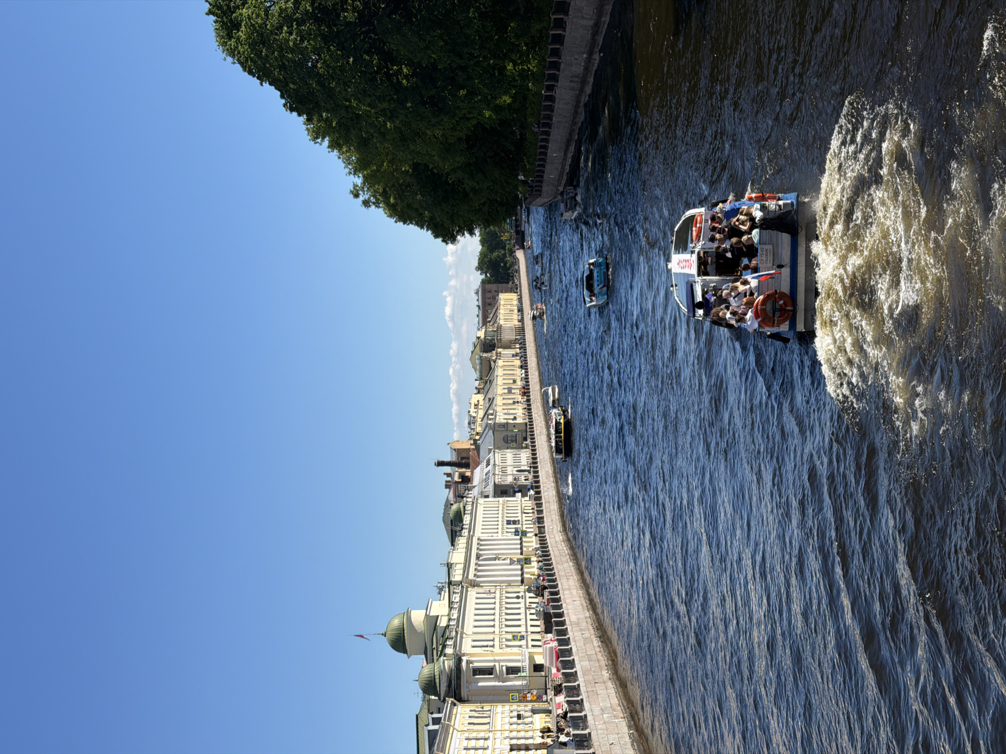 The image depicts a wide view of a vibrant canal or river in Saint Petersburg, Russia, on a clear, sunny day. The scene is dominated by the waterway, filled with various boats, and flanked by embankments.
On the left side of the frame, a continuous line of historic, neoclassical buildings stretches into the distance. These structures are primarily light yellow or cream-colored with white architectural details, including numerous columns and cornices. Several buildings feature prominent green or teal domes, with one particularly large green dome visible in the mid-left, topped by a flagpole bearing a red rectangular flag. A stone embankment, featuring a balustrade, separates the buildings from the water. A road runs along the embankment, with vehicles and pedestrians visible.
The right side of the frame features a dense, verdant line of mature trees that extend along the waterway, obscuring any structures behind them. A dark stone embankment with a simpler railing borders the river on this side.
The dark blue water of the river is rippled and choppy, reflecting the bright sunlight. Several boats navigate the waterway. In the immediate foreground, a white and blue tour boat, identified with "C3 13-19" and "РЫЖИК" (RYZHIK) on its stern, moves away from the viewer, leaving a substantial wake of white foam. The boat is crowded with passengers, mostly seated, and a red lifebuoy is attached to its rear railing. Further down the river, a light blue motorboat with a dark canopy moves towards the viewer. Multiple other smaller vessels, including at least one more tour boat, are visible in the mid-distance, moving in both directions along the canal.
The sky above is a uniform, clear blue, devoid of clouds, indicating excellent weather conditions. The strong sunlight illuminates the scene, casting defined shadows. Based on the distinctive classical architecture with domes and the characteristic canal system, the location is identified as a central canal or river in Saint Petersburg, Russia, highly likely the Fontanka River Embankment, given the specific architectural features of the buildings.