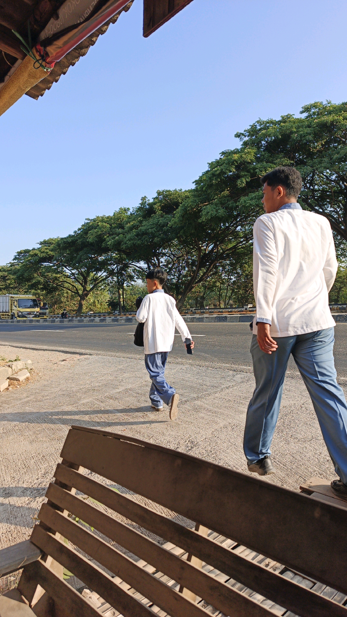 Two young men in white shirts and blue pants are walking on a sunny day along a paved path next to a road. A large truck is visible on the road in the background, as are several large trees with green foliage. A wooden bench is in the foreground of the image. The scene appears to be in Krimun, Indonesia, based on the context provided.