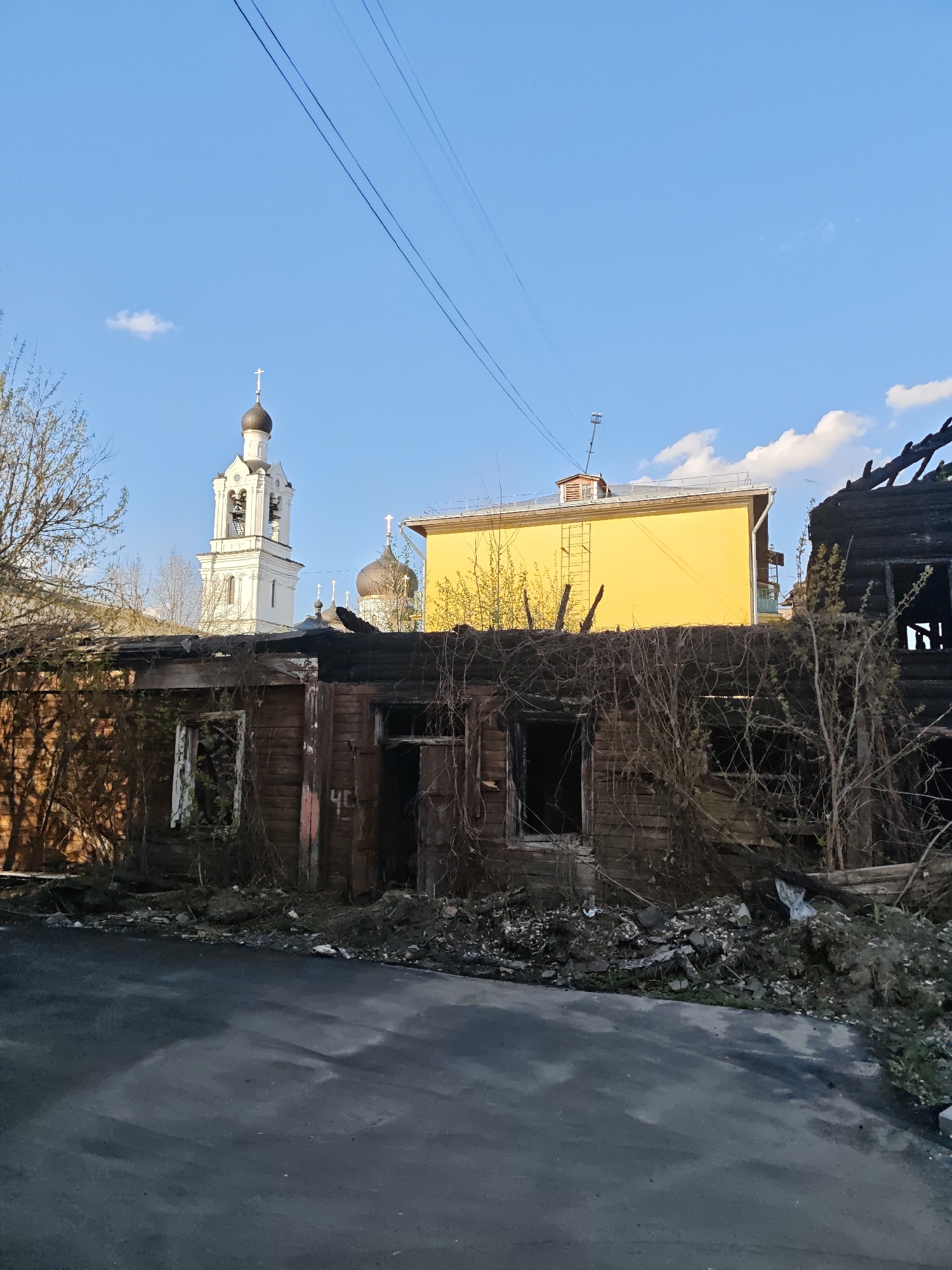 The scene depicts the aftermath of a fire in Noginsk, Russia, showing a severely damaged wooden structure with charred remains and collapsed sections. In the background are a yellow building and a white bell tower with a dark dome and cross, partially obscured by trees. The ground is covered with debris and vegetation. A partially visible number "46" is painted on a wall of the structure. Overhead, power lines stretch across the blue sky with scattered clouds. The foreground is a paved street surface.