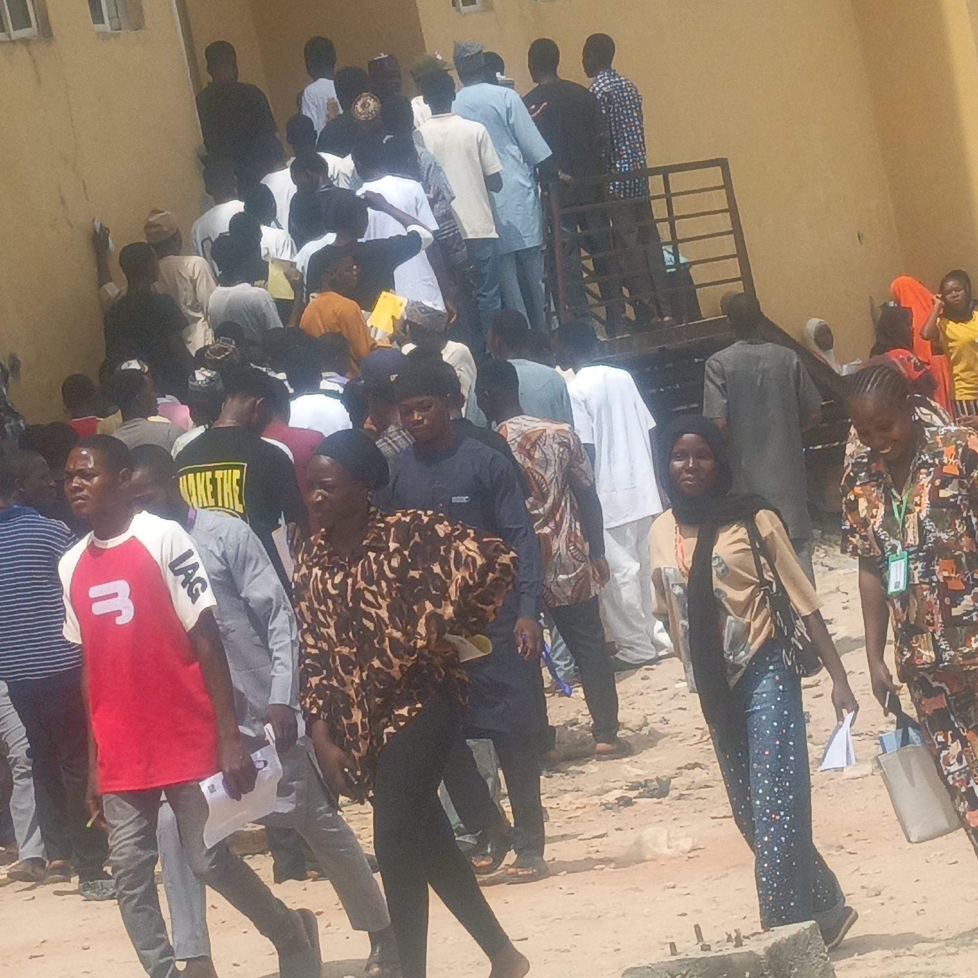 This image captures a dense gathering of many people, primarily young adults, in an outdoor setting during bright daylight. The scene appears bustling, with individuals moving and congregating near a light-colored building.
The subjects are a large crowd of diverse individuals, including both men and women, dressed in a variety of casual and semi-formal attire. Men wear t-shirts, long-sleeved shirts, and traditional tunics, while women are seen in hijabs, patterned dresses, and contemporary outfits. In the foreground, a man wears a red t-shirt with "JAG" and a white number "3" printed on it. Another person further back is seen in a black t-shirt with yellow lettering that appears to say "MAKE THE". Many individuals are holding small white papers or documents. One woman on the far right wears a patterned top and has a light green lanyard visible around her neck. Some people in the foreground look towards the camera, while others are engaged with their surroundings; a woman on the right side of the frame is smiling.
The scene is set outdoors, characterized by a dirt or dusty ground surface. A two-story building with small windows forms the backdrop on the left and right, and a black metal staircase or railing suggests access to an upper level where more people are visible. The bright, direct sunlight indicates it is daytime, likely mid-morning or afternoon, under clear weather conditions.
The activity appears to be a large public gathering, possibly involving queuing, registration, or attendance at an event, given the number of people and the documents they are holding. The crowded nature suggests an active and potentially busy process. The location cues, attire, and general atmosphere are consistent with a public area in Keffi, Nigeria.