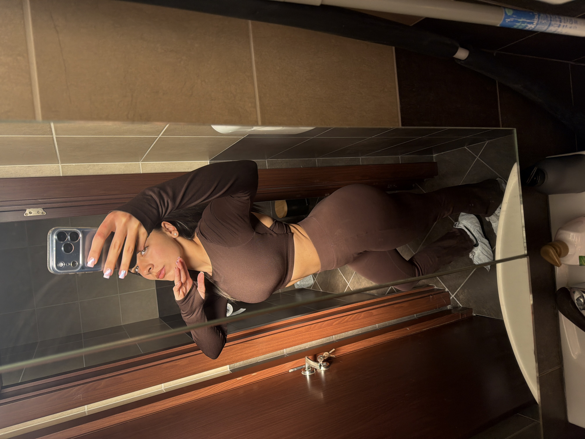 A woman is taking a selfie in a bathroom mirror. She is wearing a brown, long-sleeved crop top and brown leggings. The bathroom has brown and beige tiles. A toilet and a sink are visible in the reflection. The location is likely indoors. There is no indication of Gabrovo, Bulgaria.