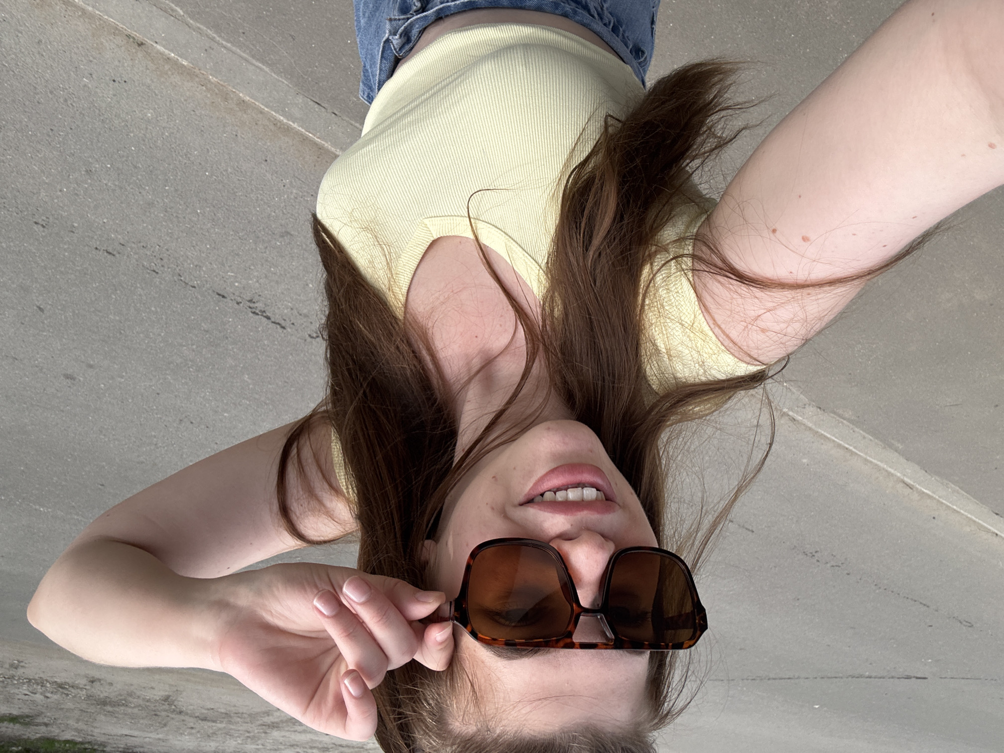 A young woman, presumably in Посёлок комбината стройматериалов-2, Russia, is holding sunglasses up to her face, upside down relative to the viewer's perspective. She is wearing a light yellow ribbed top and denim bottoms. Her hair is long and brown, with some strands obscuring her face. The background appears to be concrete.