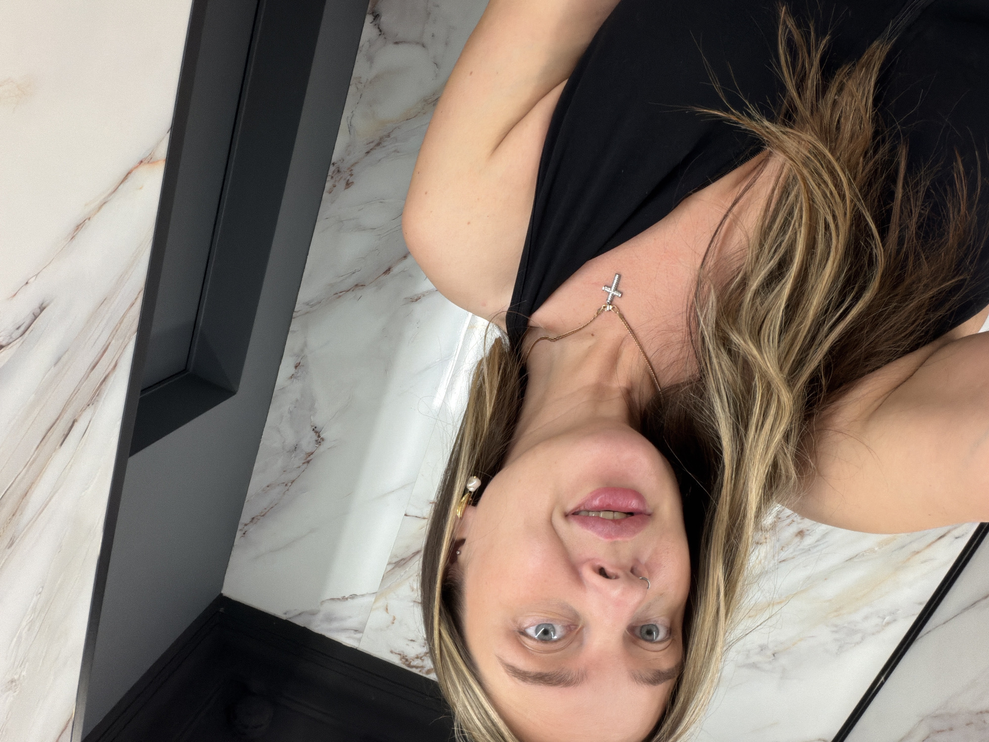 A Caucasian woman with light brown hair and blue eyes, wearing a black top and a cross necklace, is in a room with marble-patterned flooring and walls. A dark grey structure is visible on the left side of the frame. It is impossible to confirm the location in the city of Podolsk, Russia, based on the image.