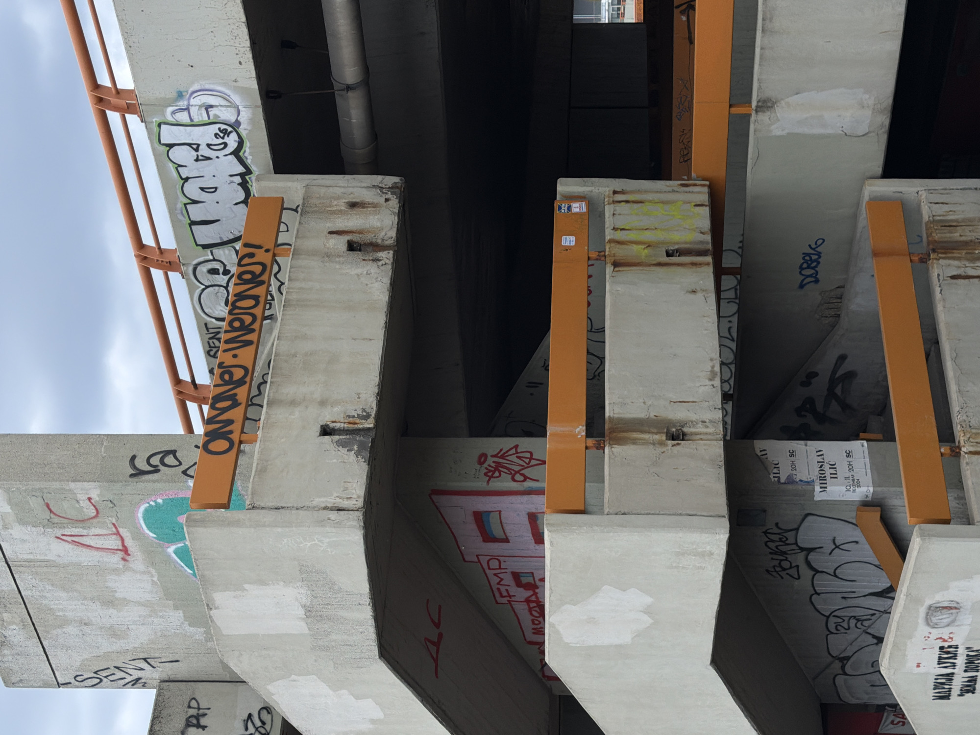 This is an image of the underside of a bridge structure in Belgrade, Serbia, as indicated by Cyrillic text on a poster and graffiti. The structure appears to be made of concrete with orange metal supports. The concrete surfaces are covered in graffiti of various styles and colors. The graffiti includes recognizable words, letters, and tags, as well as posters with Cyrillic text. Some graffiti is blurred out or faded. The environment appears to be urban, with the sky visible in the upper left corner of the image. The viewpoint is from below, looking up at the structure.