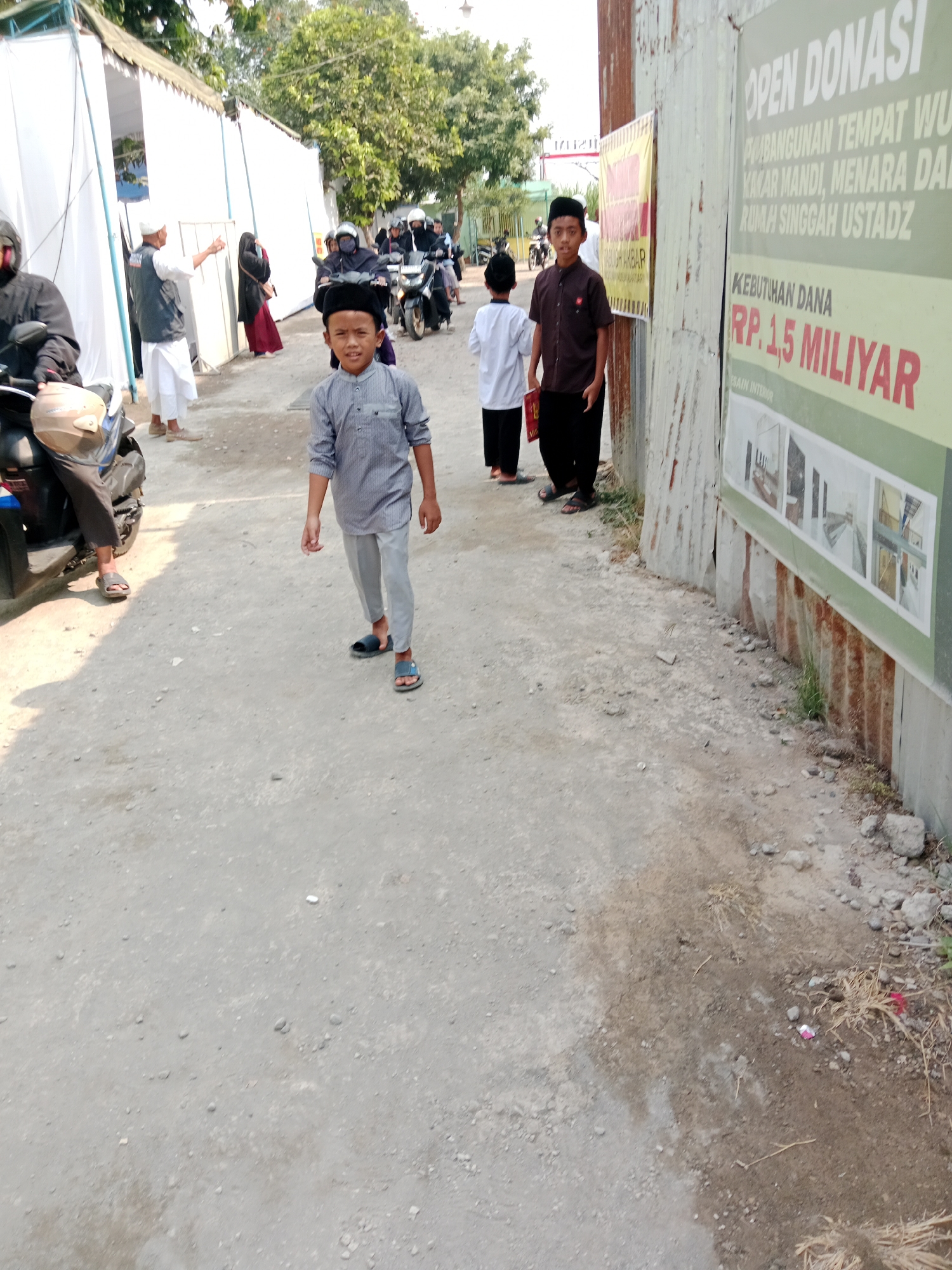 The image depicts a street scene in Kediri City, Indonesia, on a sunny day. In the foreground, a young boy in a grey striped shirt and black peci (cap) walks towards the camera. Behind him, another boy in a dark brown shirt and black pants stands next to a corrugated metal wall displaying a large green banner with text in Indonesian, including "OPEN DONASI" and "KEBUTUHAN DANA RP. 1,5 MILIYAR". Further back, several people are visible, some on motorcycles, and others walking near white tents or buildings. The ground is unpaved and dusty, with scattered rocks and dry vegetation. The overall atmosphere suggests an outdoor gathering or event, possibly a fundraising activity given the banner.