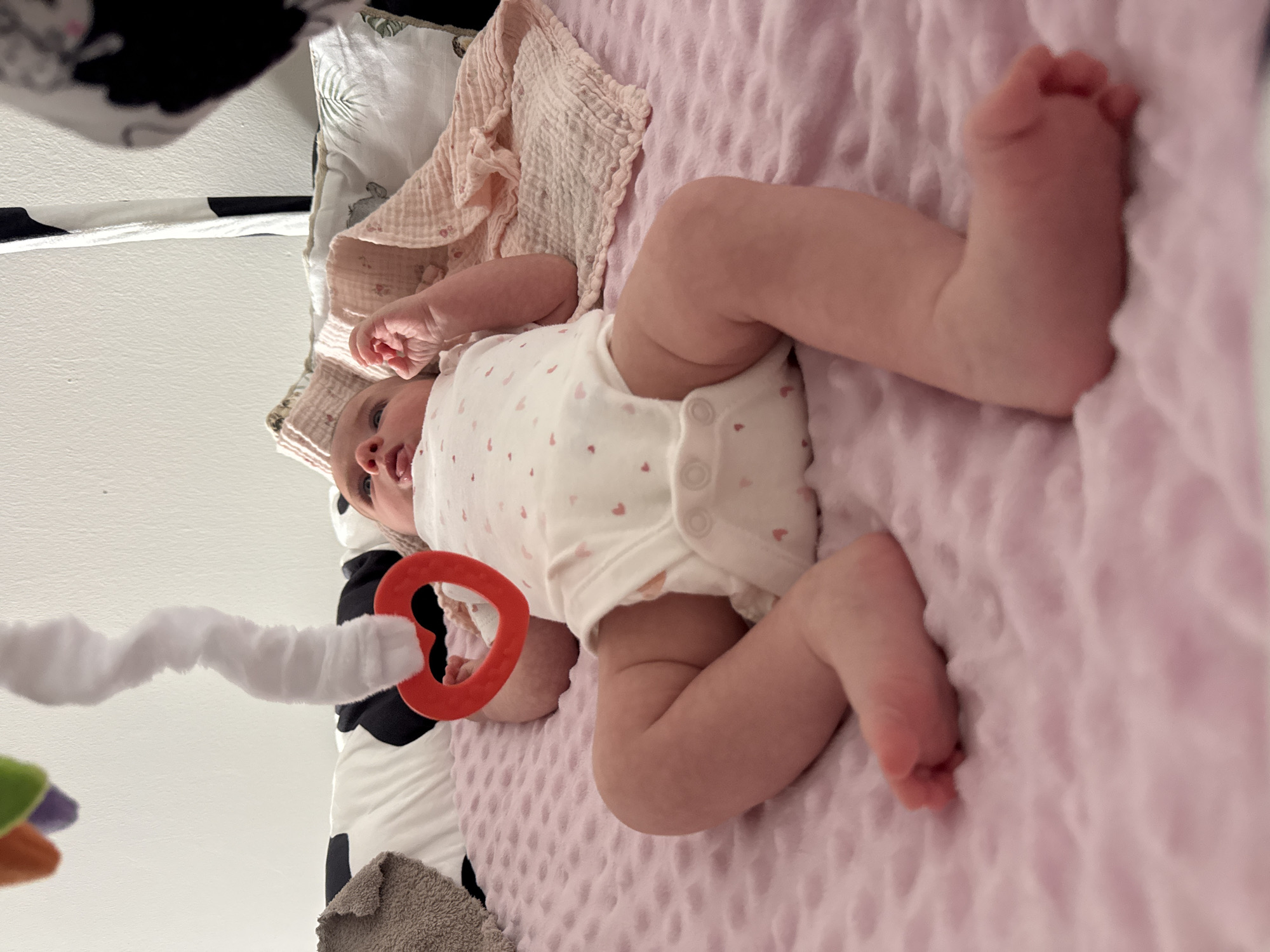 An overhead, slightly tilted shot captures a baby lying on its back on a soft, light pink textured blanket, likely indoors in a nursery or home setting. The baby is wearing a white onesie adorned with small pink heart patterns. Its head is towards the upper right of the frame, with fair skin, open eyes, and a slightly ajar mouth, looking upwards and slightly to the left.
One of the baby's hands is near a bright red, circular teething or play ring, which is attached to a white, spiral-wrapped toy arm extending from the left side of the frame. Other soft, colorful toys (green, purple, orange) are visible but out of focus further to the left. Near the baby's head, a crinkled, light pink muslin-like blanket is draped, and a pillow or patterned fabric with a subtle design is visible beneath it. The baby's bare feet are curled and resting on the pink blanket towards the lower right. A black and white patterned fabric is visible along the far left edge of the image. The scene suggests a calm, playful moment, likely during the daytime. No text is visible in the image. While the provided context is Isola delle Femmine, Italy, no specific geographical cues are visible within the image itself.