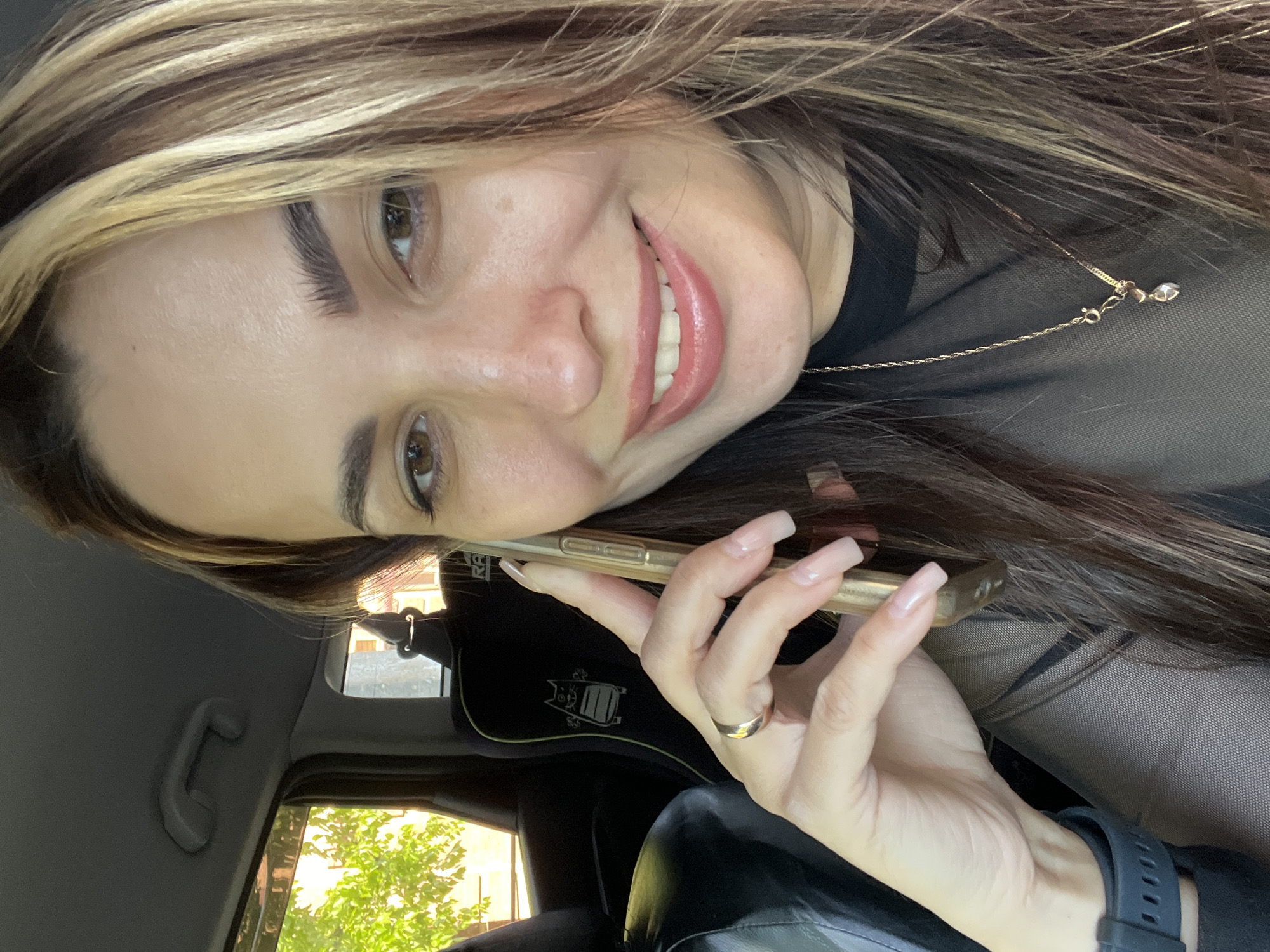 A woman with brown and blonde hair is in a car in an unknown city, holding a gold iPhone against her left ear. She is wearing a black mesh top and a gold necklace with a pendant. She has long, square-shaped manicured nails and a gold ring on her finger. A black watch is visible on her left wrist. The interior of the car includes a black headrest with a logo, a grab handle, and a view of a building with green trees through the window.