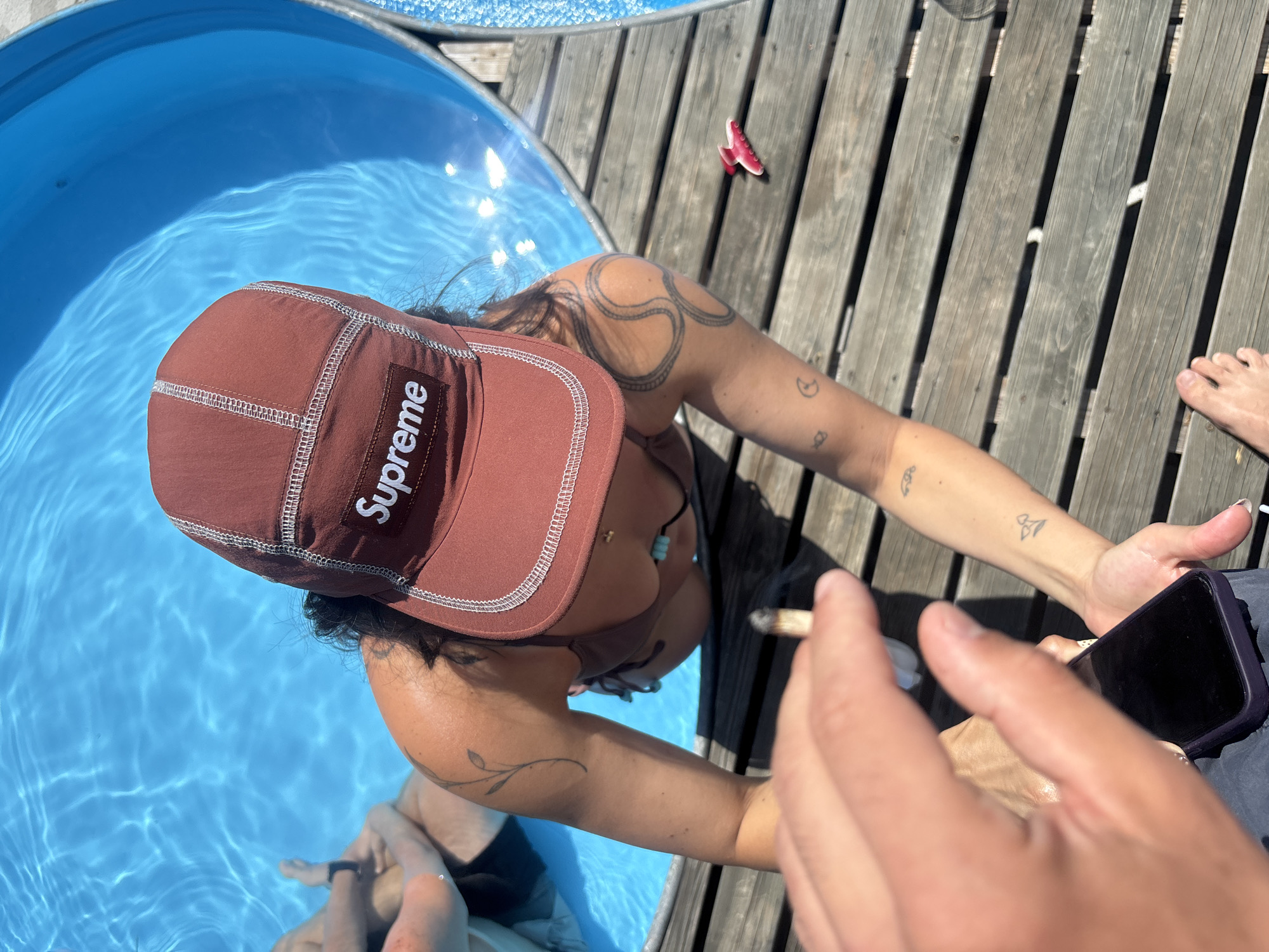 A person wearing a brown Supreme baseball cap and a brown bikini top is leaning into a circular blue pool filled with clear water. They have several tattoos on their arms, including a plant-like design on their left upper arm and a spiral design on their right upper arm. Another person, partially visible, is in the pool with them. The pool is surrounded by a wooden deck. A hand holding a smartphone is in the foreground, obscuring part of the scene. A pink hair clip is visible on the deck to the right of the pool. The overall setting appears to be an outdoor recreational area, possibly a backyard or a poolside area. The image does not provide any information to ascertain its location in San Lorenzo De Tormes, Spain.