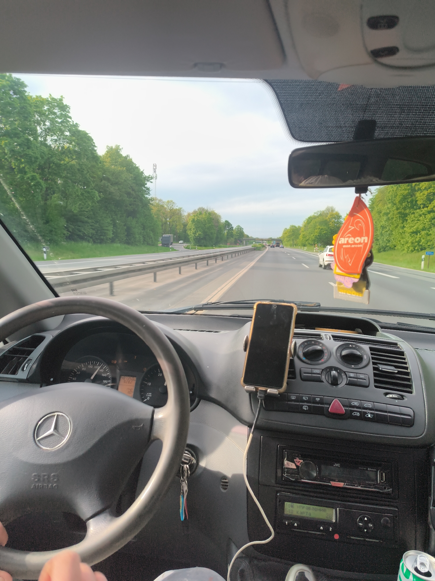 The image offers a first-person view from inside a Mercedes-Benz vehicle, capturing a drive on a multi-lane highway near Ascheberg, Germany. The scene depicts a late afternoon or early evening setting, with soft natural light under a partly cloudy sky.
In the foreground, the driver's hands are visible on the steering wheel, which bears the Mercedes-Benz logo and "SRS AIRBAG" text. The dashboard includes an instrument cluster showing the car traveling at approximately 110-120 km/h. The odometer reads 187,350 km, with a trip meter showing 926.2 km, and the digital clock displays 17:42.
A smartphone in a light-colored case is mounted on the central console, connected by a charging cable. Below it, the car's infotainment system features a JVC brand radio and a lower display unit showing Cyrillic text: "Управление" (Control) and "32 карта" (32 map), suggesting a navigation or multimedia interface. Car keys dangle from the ignition. A can, possibly a soft drink, is partially visible in the bottom right corner. An orange "areon" branded air freshener hangs from the rearview mirror, which reflects a portion of the driver's upper body.
Outside the car, the highway stretches into the distance, lined with green trees. Other vehicles, including a white car and a distant truck, are visible ahead. A metal guardrail separates the highway lanes from the surrounding landscape. The overall impression is that of a calm, routine drive on a European autobahn.