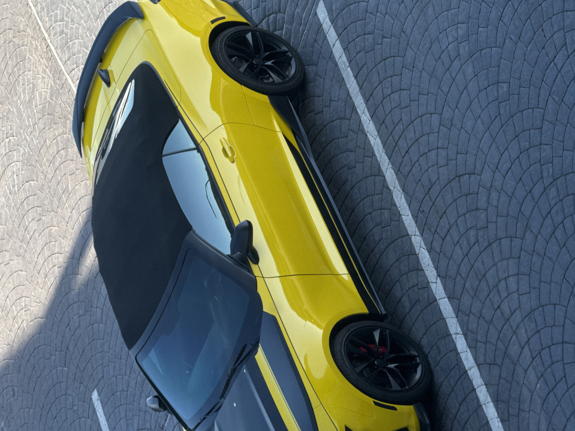 A yellow convertible Chevrolet Camaro with black racing stripes parked on a cobblestone surface marked with white lines, possibly in a parking lot. The car's black soft top is up. The scene is shot from a high angle, looking down on the car. The environment appears to be outdoors, with sunlight casting shadows on the ground. The image does not provide any specific information or visual cues to suggest the location is Sochi, Russia.