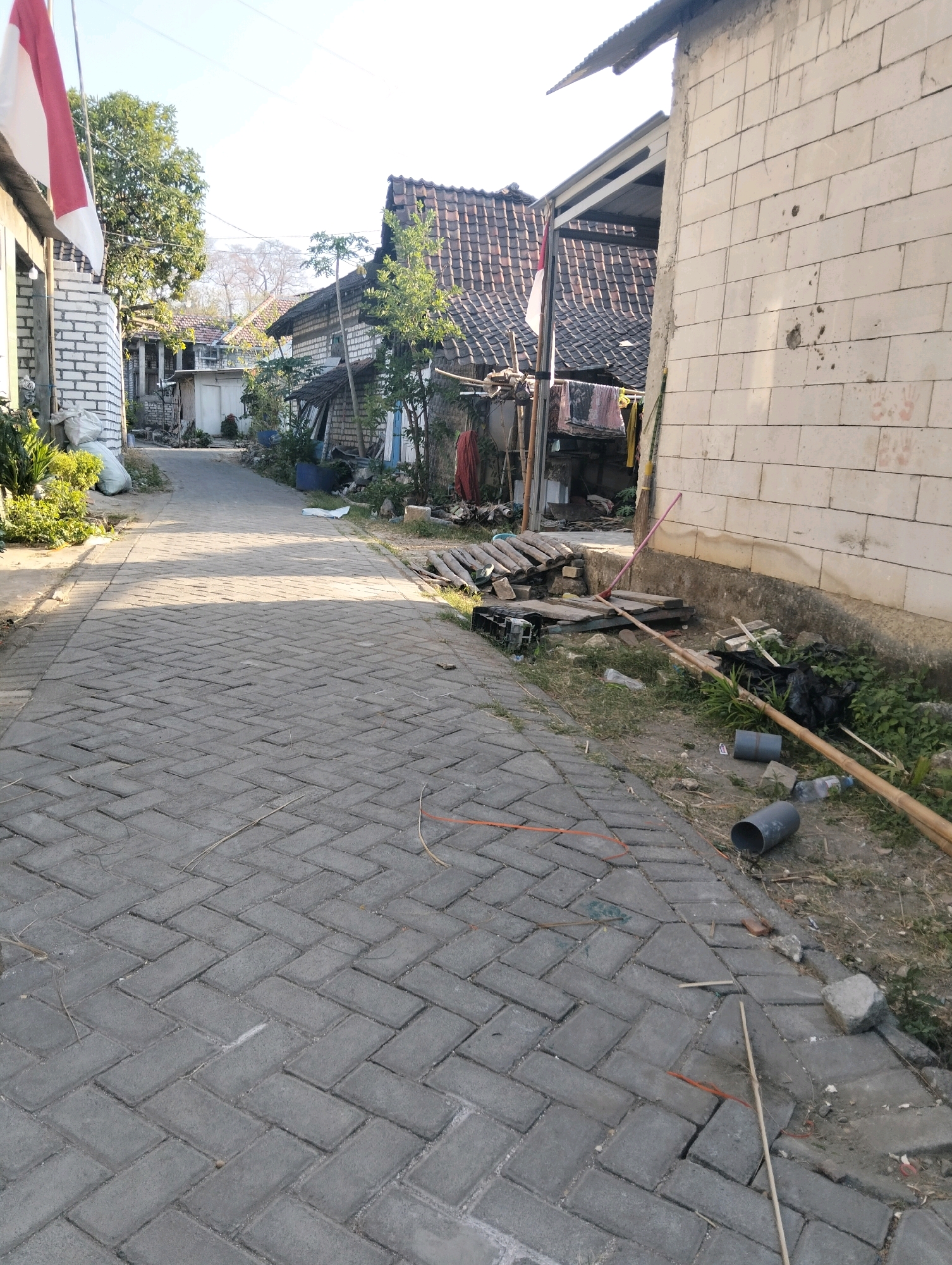 The image depicts a narrow, paved alleyway in a residential area, likely in Drajat, Indonesia. The alley is paved with interlocking grey bricks laid in a herringbone pattern. To the left, a white brick wall is visible, topped with a corrugated metal roof and adorned with a red and white Indonesian flag. Lush green foliage grows near the base of this wall. Further down the alley, several houses with tiled roofs are visible, interspersed with trees and more greenery. The right side of the alley is bordered by a rough, light-colored brick wall. The ground beside this wall is cluttered with debris, including a pink broom, several pieces of bamboo, a black plastic bag, gray PVC pipes, and some scattered trash. The scene is illuminated by natural sunlight, casting shadows across the paved surface. The sky above is clear and blue.