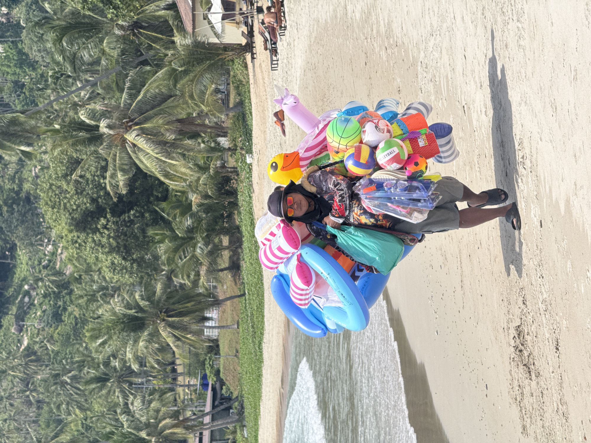 A person walks along a sandy beach on Ko Samui, Thailand, carrying a large assortment of colorful inflatable items and beach balls. The person wears sunglasses, a hat, and shorts, and has a green bag slung over their shoulder. The beach is lined with palm trees, and lounge chairs and umbrellas are visible in the background. The ocean waves are gently breaking on the shore.