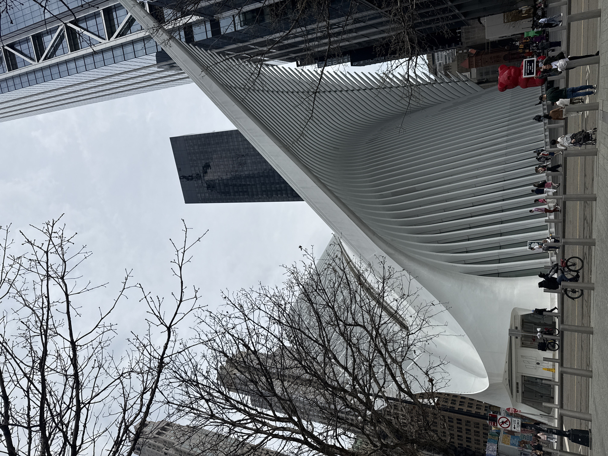 The image shows a modern architectural structure in New York City. The white, ribbed building is the Oculus at the World Trade Center transportation hub. Several people are walking and cycling in the foreground along a pedestrian walkway beside the building. In the background are skyscrapers, some appearing to be under construction or covered. Bare trees are visible in the lower part of the frame. The sky is overcast.