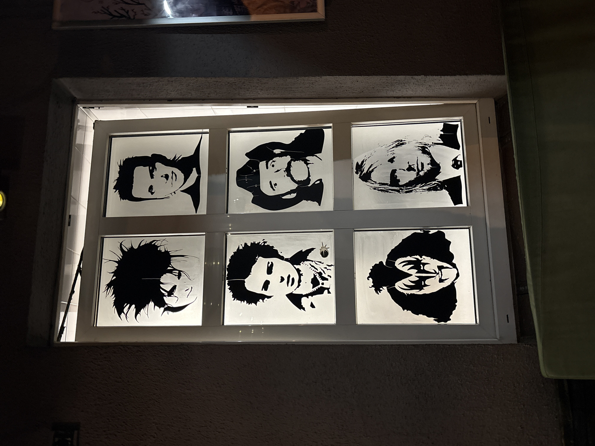 Six portraits are displayed in a white-framed window. The portraits are black silhouettes on white backgrounds, depicting people. The window is set into a textured, dark wall. A portion of a green wall or surface is visible to the right of the window. The location is Minsk, Belarus.