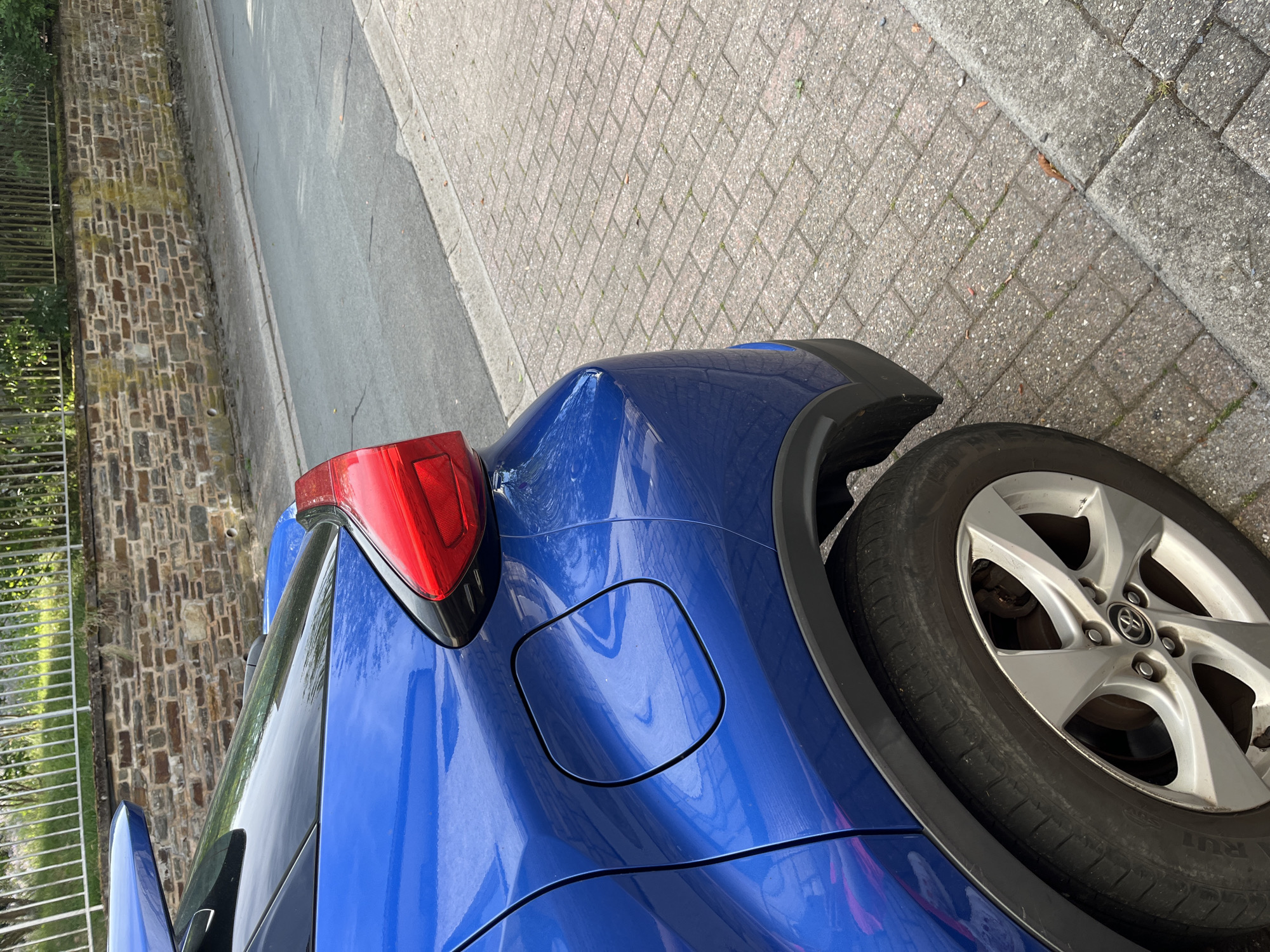 The image shows the rear driver's side of a blue car parked on a cobblestone sidewalk. The car has a damaged rear bumper and taillight. The background includes a stone wall, a street, and a fence. The location is Saint-Vith, Belgium. The scene appears to be a typical street view in a European town. No people are visible in the image.