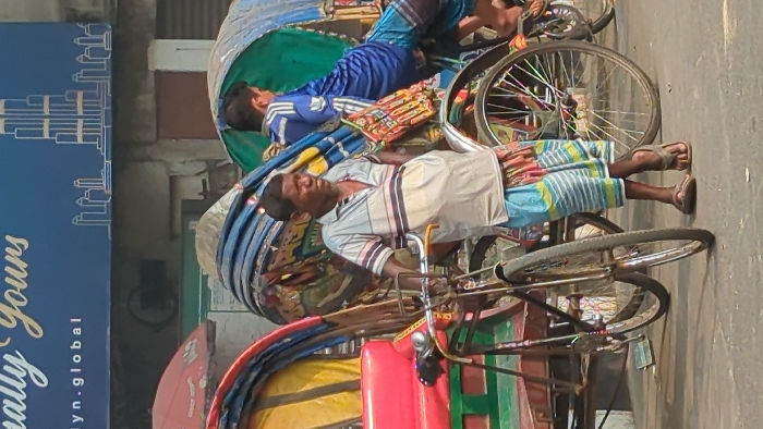 The image depicts a daytime street scene in Dhaka, Bangladesh, centered on cycle rickshaws and individuals.
In the foreground, an adult male, identified as a rickshaw driver, stands upright, resting his right hand on the handlebars of a cycle rickshaw. He wears a light-colored, horizontally striped polo shirt, a multi-patterned blue, green, and orange lungi, and brown flip-flops. Perspiration is visible on his shirt. His rickshaw features a blue and multi-colored patterned fabric canopy. A small, intricately woven, colorful bag hangs from the front of this rickshaw's passenger compartment.
To the right of the primary subject, a younger male is seated or leaning in the passenger compartment of a second cycle rickshaw, facing away from the camera. He wears a blue long-sleeved athletic shirt with white stripes and blue patterned shorts. This rickshaw has a green fabric canopy.
To the left, a third cycle rickshaw is partially visible, featuring a bright red passenger seat, green side panels, and a yellow and blue striped canopy. The ground is a paved, dusty surface.
In the background, a building features a dark blue sign with white text, partially visible as "Finally Yours" and "lyn.global". The sign also displays a white line-art illustration of a cityscape, including a prominent, stylized skyscraper.