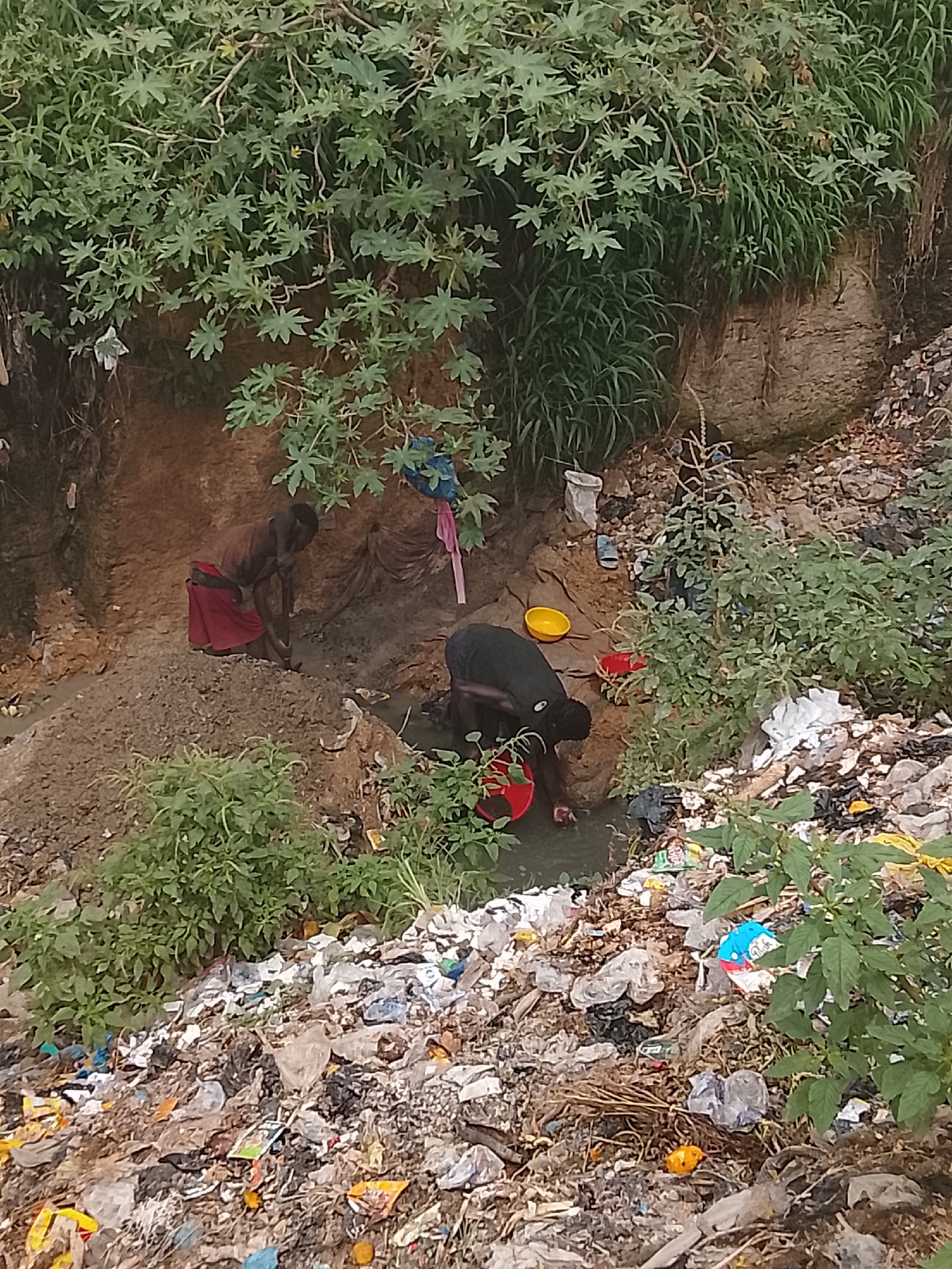 In Gwong, Nigeria, a scene unfolds by a polluted ditch, where two individuals, one wearing a red cloth around their waist and a brown top, and another in a black shirt, are engaged in washing. The environment is characterized by a steep, eroded embankment covered in lush green foliage. The ditch itself is a waterway, murky and shallow, serving as a washing area. Scattered around are remnants of human activity, including plastic bottles, wrappers, and other assorted waste, testament to the pervasive presence of litter. A yellow bowl and a red bucket are present near the washing area, likely used for carrying water. The ground is a mixture of dirt, sand, and debris, with clumps of dry grass and twigs adding to the disarray. The overall impression is one of a functional but neglected space, where daily chores are performed amidst an environment burdened by waste.