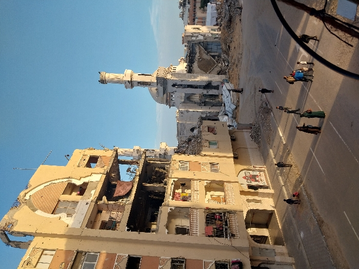 The image depicts a scene of extensive urban destruction in Khan Yunis, Palestinian Territory, under a clear blue sky. On the left, a multi-story, light-colored building is severely damaged, with multiple floors exhibiting exposed interiors, shattered windows, and structural collapse, including a large arched opening on its upper facade. Clothes are hanging from several balconies on the intact sections. To the right, in the midground, the remnants of a large, light-colored mosque are visible, featuring a partially intact dome and a damaged minaret, surrounded by significant rubble and debris.
On the street level below, an asphalt road with white markings is visible. Approximately 15 individuals, including adults and children, are present on the street and sidewalk. Several people are walking, while others are standing in small clusters. One person is bending down near a child on the sidewalk. The area between the street and the mosque consists of substantial piles of debris and dirt. A dark, curved metal object, possibly a railing, is partially visible in the bottom right foreground.