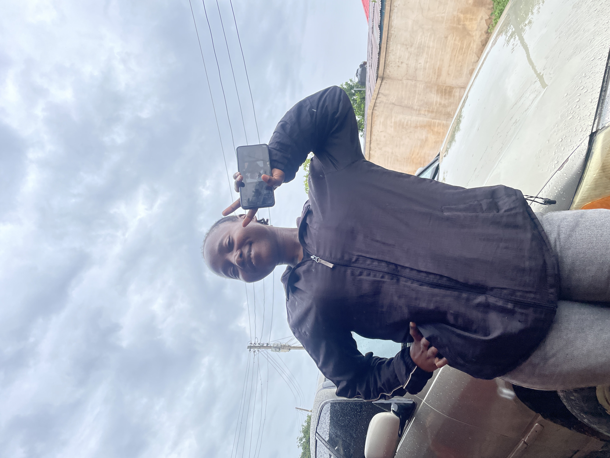 A woman stands next to a parked car under an overcast sky in Jos, Nigeria. She is angled towards the camera and holds a smartphone in her left hand, displaying an image of herself. Her right hand is gesturing a peace sign. She wears a dark-colored jacket over gray pants. The backdrop includes power lines and a building. Rain appears to be present, evident on the car's surface.
