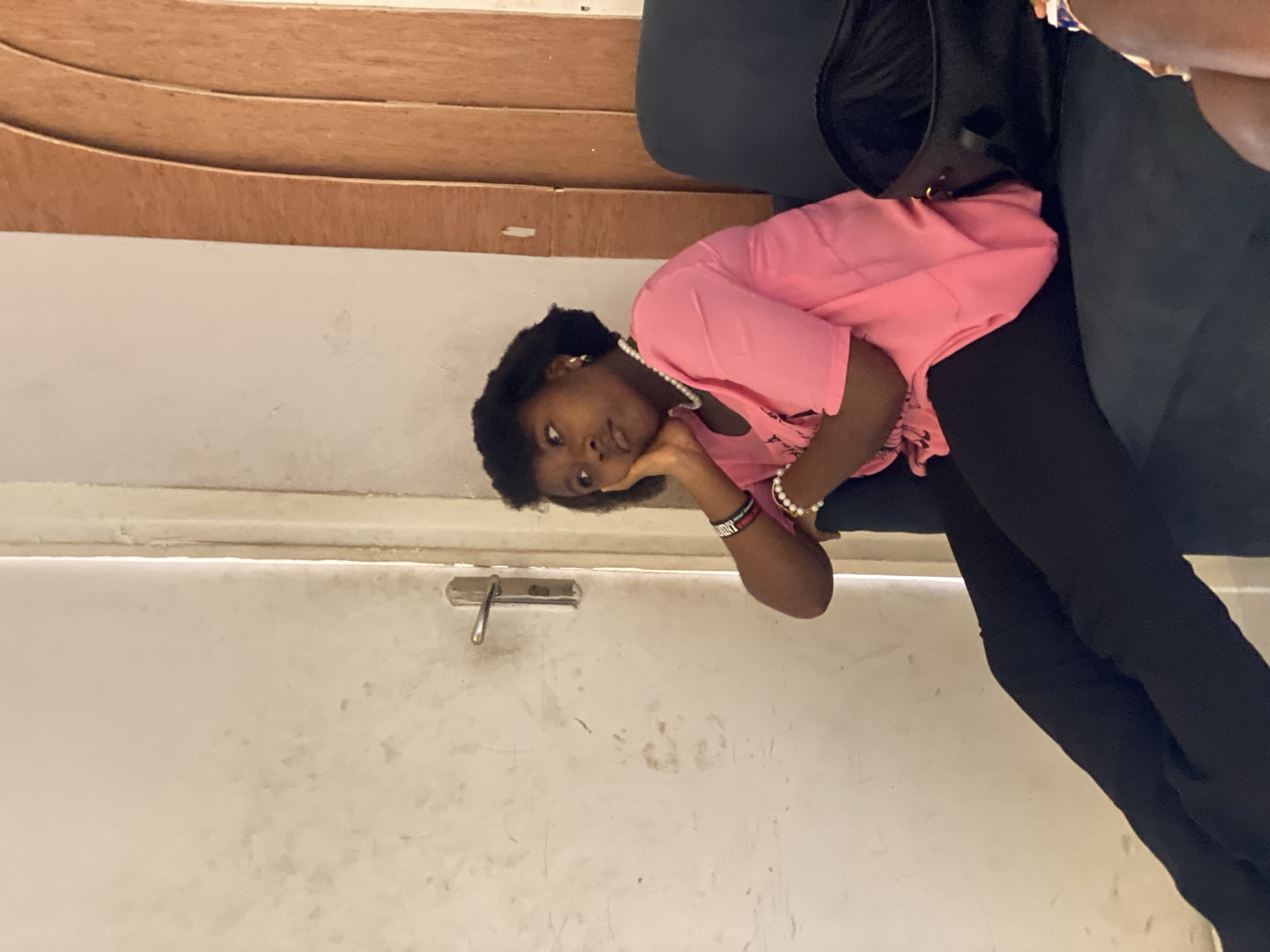 A young woman rests her head on her hand, appearing contemplative. She wears a pink shirt, black pants, and a pearl necklace. A black bag sits next to her. The background includes a white wall with a door handle and a wooden structure above it. The location is unspecified, and there's no information in the image to connect it to the city of Badagry, Nigeria.