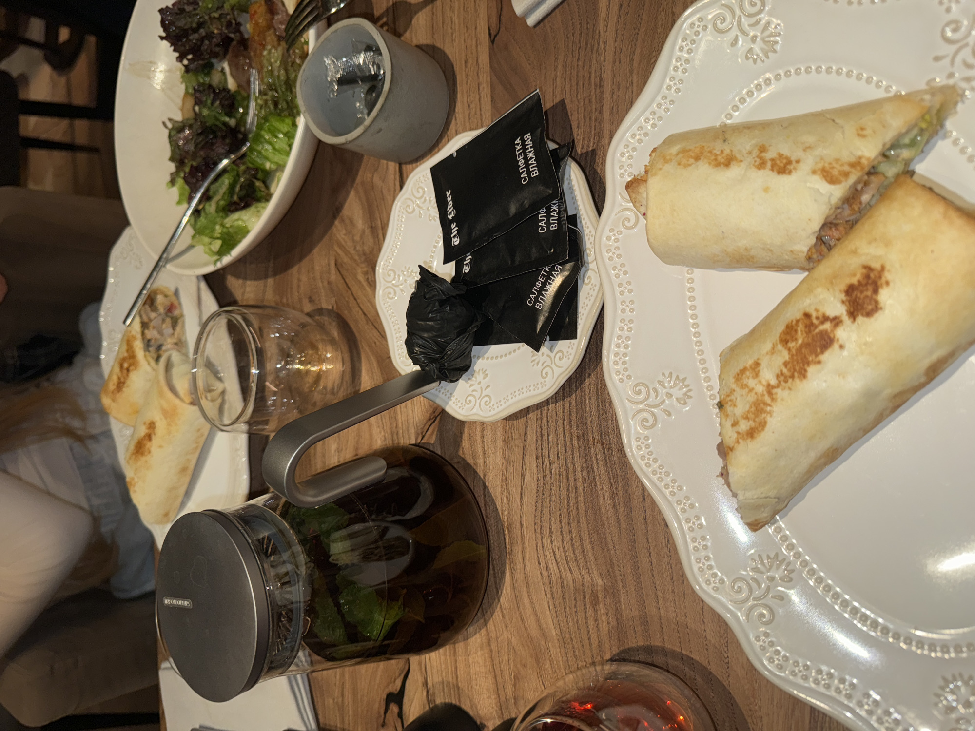 The image depicts a meal on a wooden table, likely in a restaurant. A plate of what appears to be a wrap or burrito sits on a decorative white plate. Beside it, a smaller decorative plate holds black packets with Russian text ("САЛФЕТКА ВЛАЖНАЯ"), along with a crumpled black object. A tall grey cup, a glass, and a clear teapot with leaves inside are also on the table. Additionally, there is a partially visible salad and a smaller wrap or similar food on a separate white plate to the left. The location cannot be definitively determined as Moscow, Russia, from the visual evidence alone.