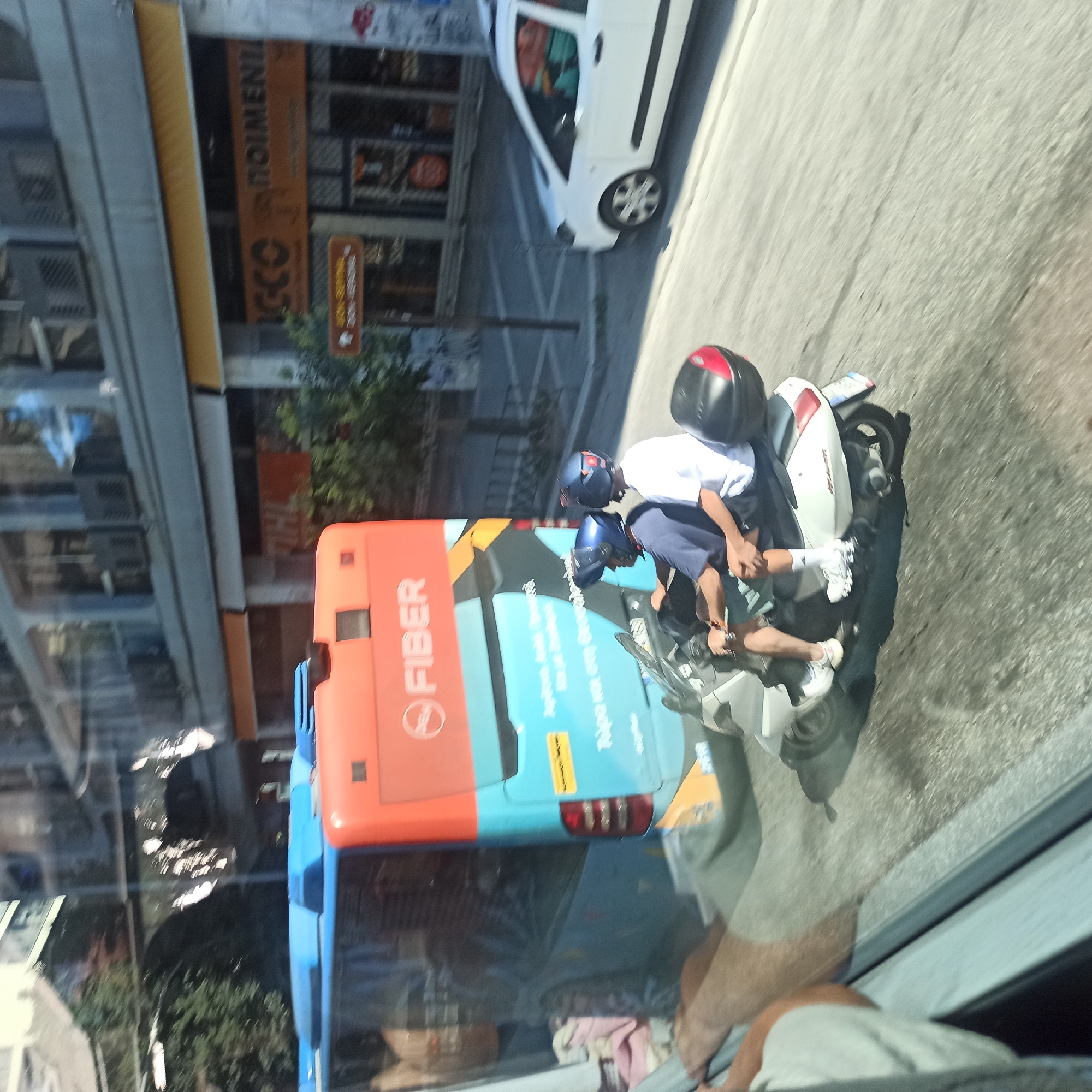 The image captures a street scene in Thessaloniki, Greece, with a focus on two individuals riding a scooter. A blue and orange bus is prominently featured in the background. The scooter, a white Honda, is moving from left to right, with the rider in front wearing a dark blue t-shirt and helmet, and the passenger behind wearing a white t-shirt and a dark helmet. Both are wearing helmets, and the passenger has a black storage box on the back of the scooter. The bus behind them displays the "FIBER" logo and has Greek text on its side. In the further background, buildings line the street, with awnings visible over shop entrances. A white car is parked on the right side of the road. The scene appears to be a sunlit day, with reflections visible on the bus windows. The road surface is asphalt with some visible tire marks.