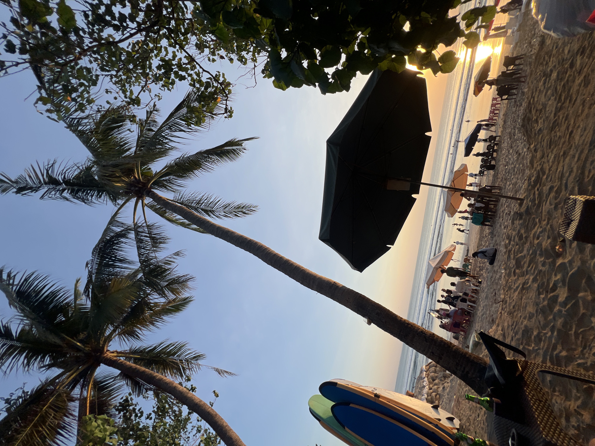 The image captures a beach scene in Kuta, Indonesia, at sunset. The foreground features a table with two green bottles, possibly beer, on a sandy surface, with a large black umbrella casting a shadow over it. To the left, several surfboards are propped up, their colorful designs visible. In the middle ground, people are seated on chairs and the sand under various umbrellas, some appearing to be enjoying a meal or drinks. Further back, the ocean stretches to the horizon, with people visible in the water and on the shore. Two palm trees lean towards the sky, their fronds silhouetted against the clear blue sky, which transitions to a warm orange hue near the horizon, indicative of the setting sun. The overall atmosphere suggests a relaxed evening at a popular tourist destination.