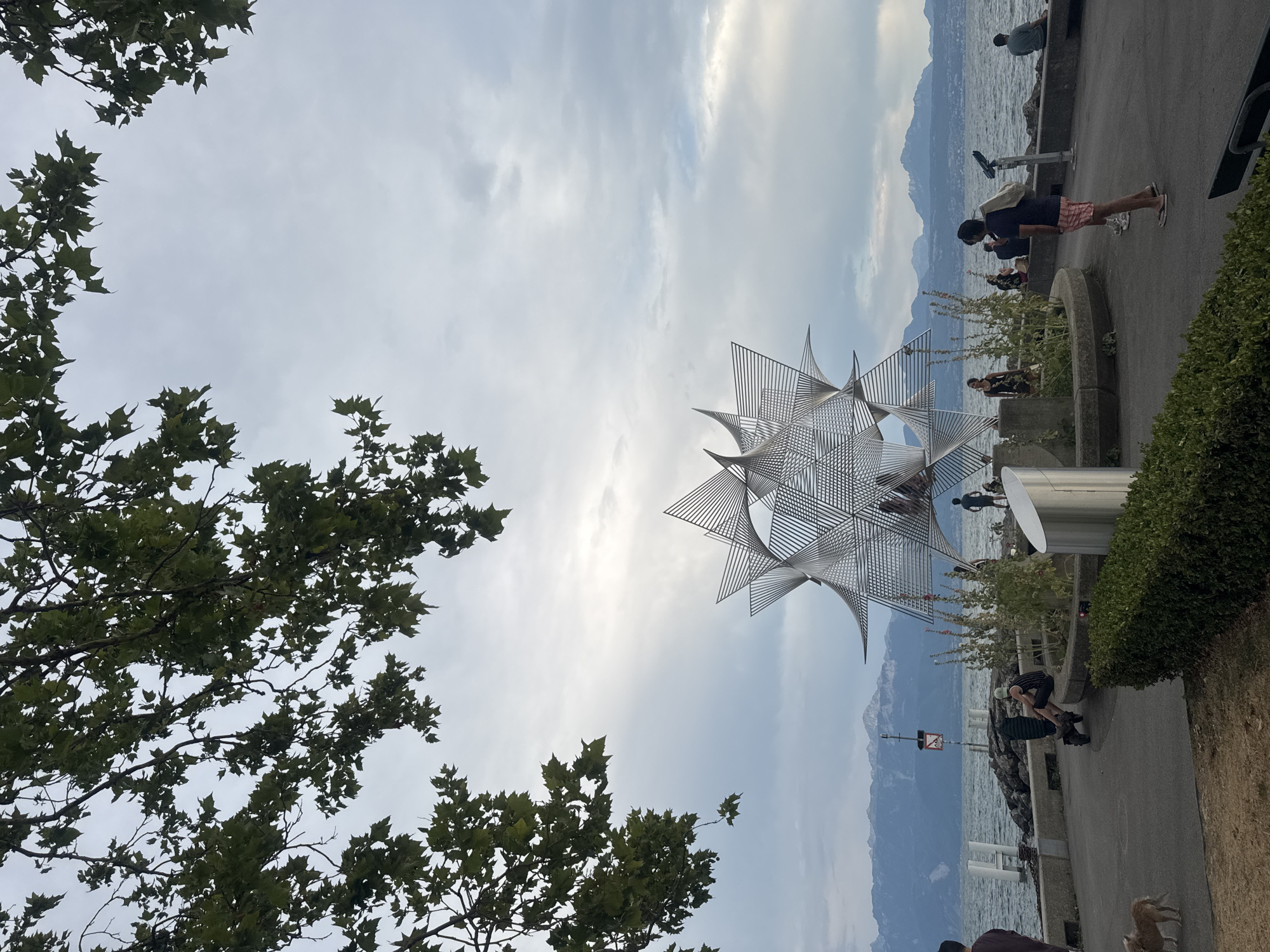The image depicts a scenic view of Ouchy, a district and former municipality in the city of Lausanne, Switzerland, on a cloudy day. In the foreground, a large, intricate metal sculpture resembling a starburst or a geodesic dome stands prominently. The sculpture is composed of numerous thin, metallic strands that create a complex geometric pattern. Surrounding the base of the sculpture is a circular garden area with low-lying plants.
A paved walkway runs along the waterfront, with several people present. To the left, a person with dark hair is partially visible, and a small tan dog is walking on the grass. Further along the walkway, a person sits on a scooter. Several other individuals are seated on a low wall overlooking the water, some appearing to converse or observe the surroundings. One person stands near a telescope, looking out at the lake.
The lake is visible in the middle ground, with calm water reflecting the overcast sky. In the distance, a mountain range, likely the Alps, forms a majestic backdrop, with its peaks partially obscured by clouds. The shoreline is lined with large rocks and a concrete barrier.
Overhanging the scene from the top of the image are the branches and leaves of a large tree, adding a natural frame to the composition. The overall atmosphere is serene, with a blend of modern art, natural landscape, and human activity by the lakeside.