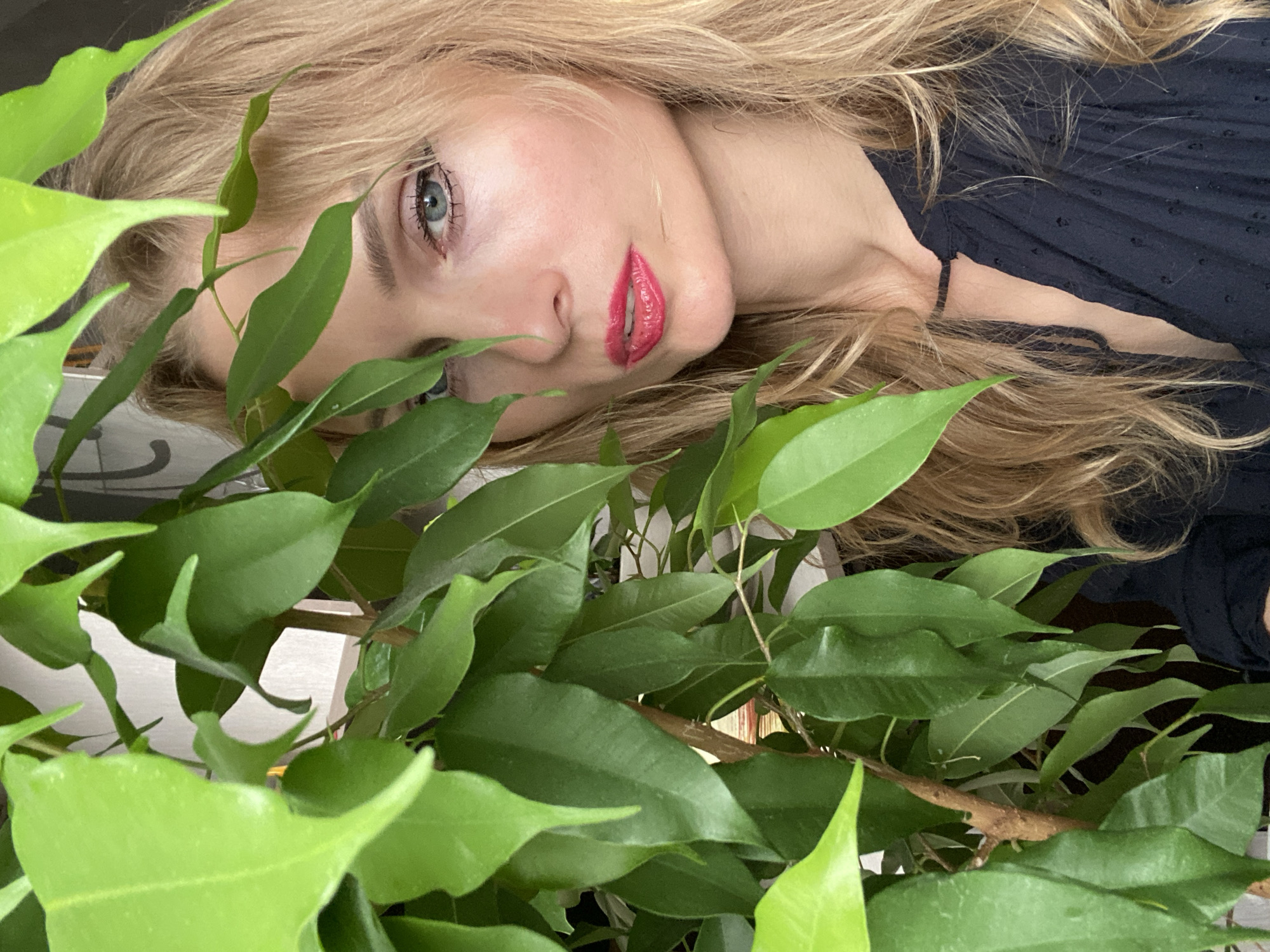 A medium shot, upper body portrait captures a fair-skinned woman with blonde hair and bright blue eyes, partially obscured by the vibrant green leaves of a ficus plant. She is looking directly at the camera, her lips painted a glossy, bright pink. The background is indistinct, suggesting an indoor setting, possibly a home in Lida, Belarus, where the natural light illuminates her face. She is wearing a dark navy, loose-fitting top with a subtle textured pattern and a keyhole neckline. The image is sharp, focusing on the woman's face and the surrounding foliage.