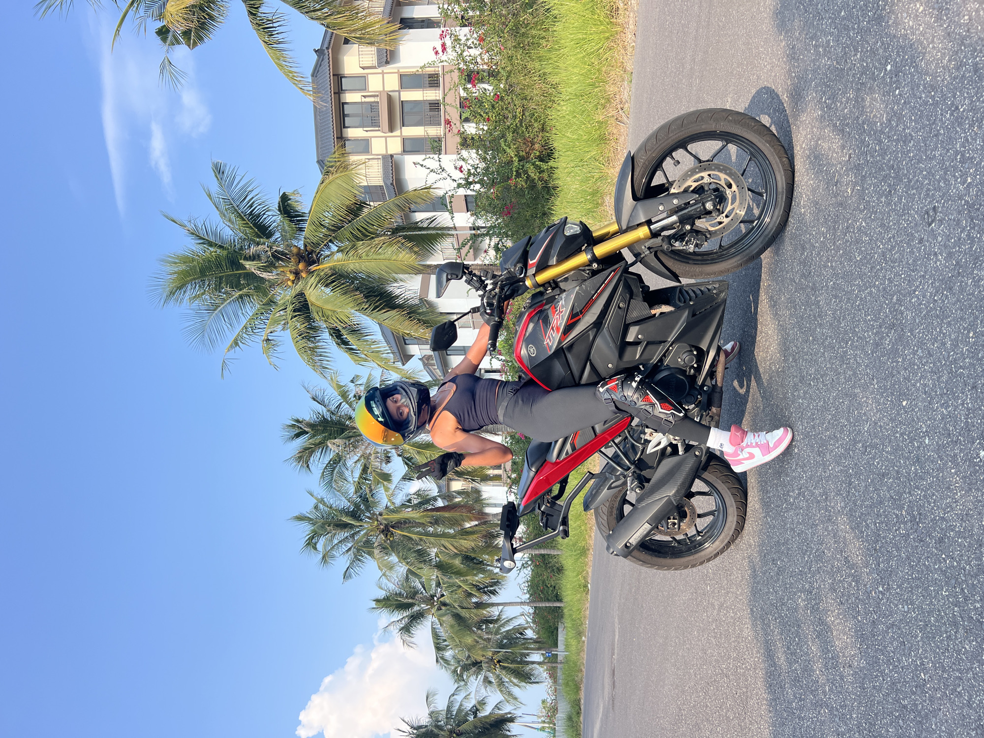 A person is sitting on a motorcycle parked on a paved road. The person wears a helmet, tank top, leggings, and pink sneakers. Palm trees line the roadside and a building is visible in the background. The scene is situated in the Nam Nha Trang Ward, Vietnam.