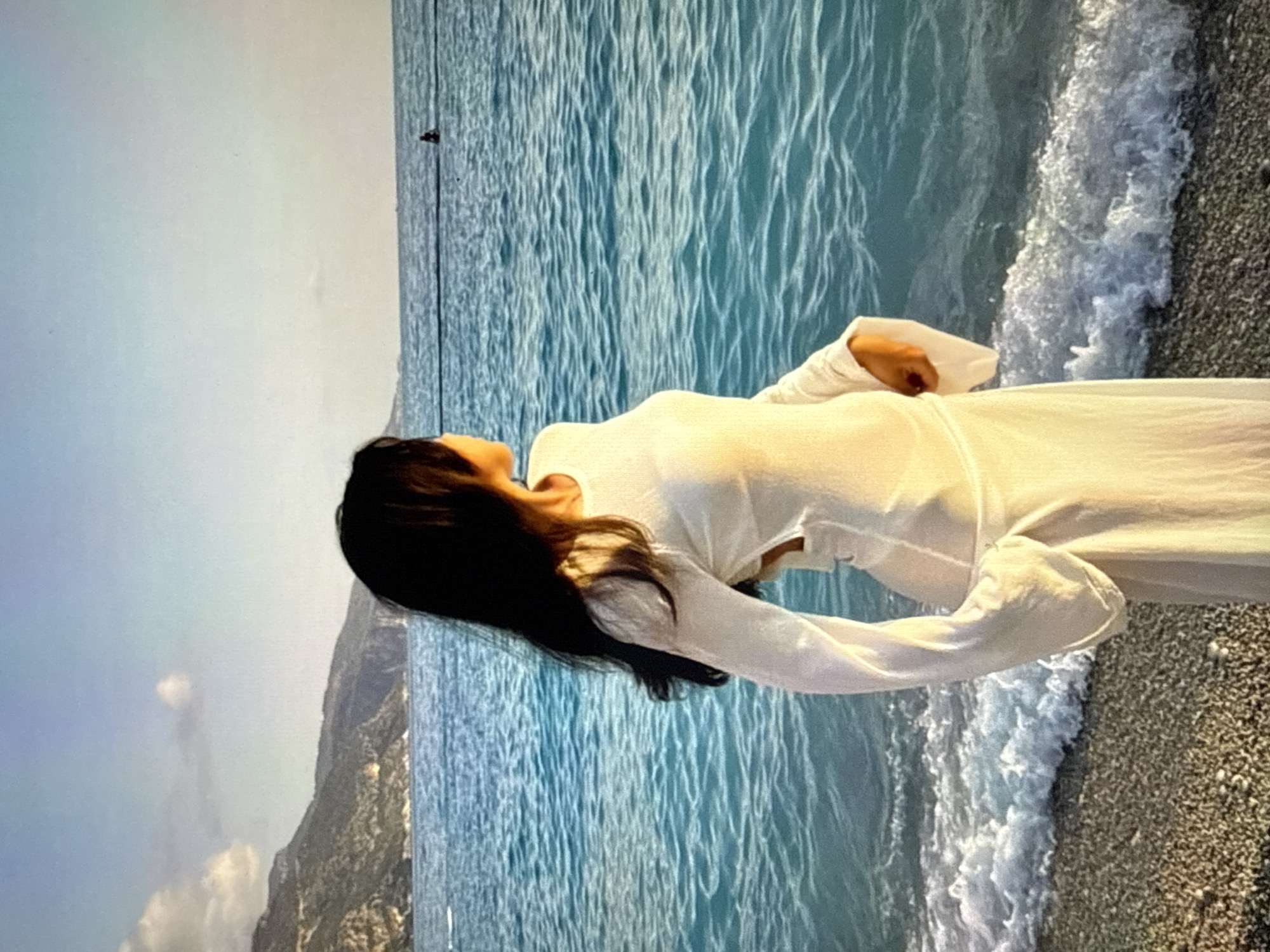 A woman with long, dark hair stands on a pebble beach, facing away from the camera and looking out at the turquoise ocean. She is wearing a long, white, flowing dress with flared sleeves. The dress has cutouts on the sides of the torso. Waves are gently breaking on the shore in front of her. In the distance, the ocean stretches to the horizon, and a faint silhouette of a person can be seen in the water. Mountains are visible in the background, and the sky is a light blue with a few wispy clouds. The scene appears to be a coastal location, possibly a beach resort. The specific location in Găvănoasa, Moldova is not discernible from the image alone.