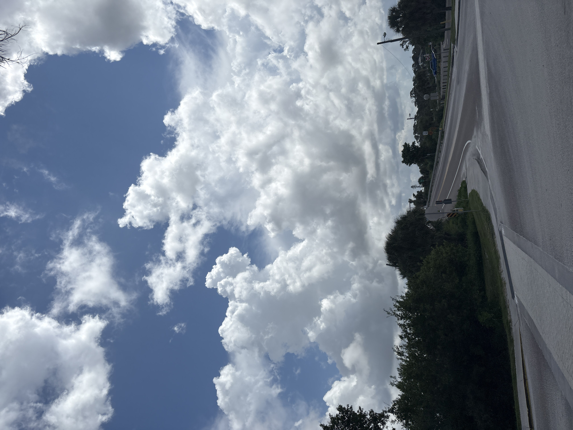 The image captures an intersection in Englewood, United States, under a dramatic sky filled with billowing cumulus clouds. The foreground features a paved road and sidewalk, with a crosswalk marked by white stripes. To the left, lush green foliage and trees line the roadside. The road curves towards the right, leading towards a distant landscape dotted with more trees and a few buildings. Traffic signals and streetlights are visible along the road. A blue vehicle is parked on the right side of the road in the distance. The overall scene suggests a partly cloudy day in a suburban or semi-rural area.