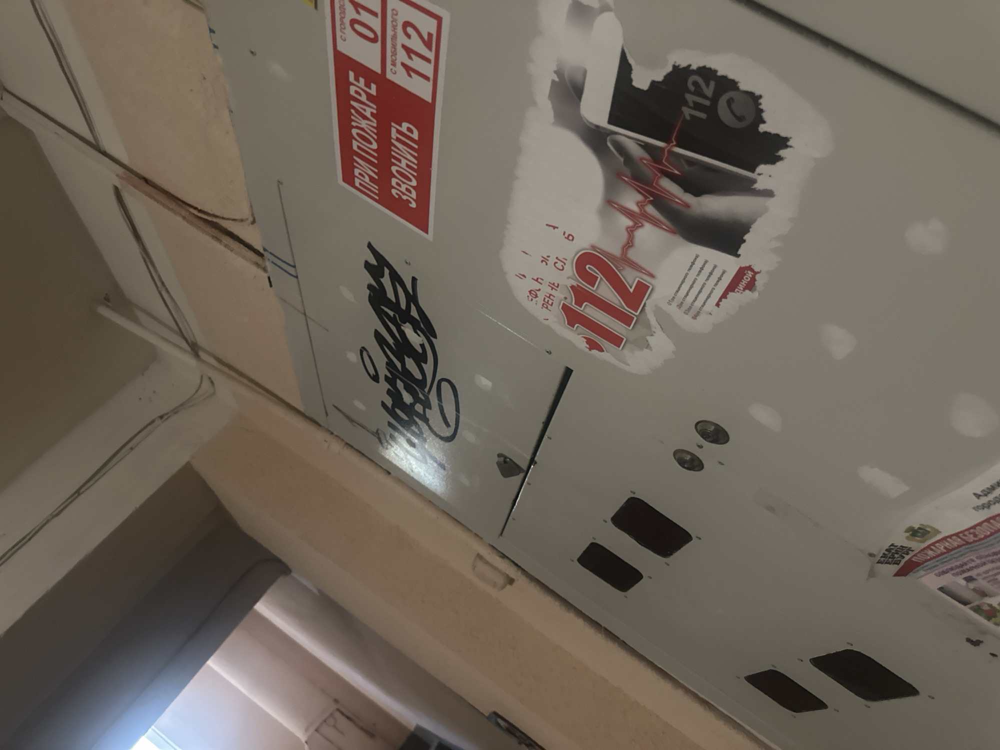 This media file captures an interior scene, likely a hallway or a wall in a public building, in Yekaterinburg, Russia. The primary focus is on a white metal surface adorned with various stickers and graffiti.
Prominently displayed is a red sticker with Russian text that translates to "In case of fire, call 01" and "From a mobile phone, call 112". Adjacent to this is another sticker featuring the number "112" in bold red, superimposed on an image of a hand holding a smartphone with a medical-like waveform on the screen. The graphic implies an emergency service, possibly for fire or ambulance. Further down, a smaller sticker contains text with "112" again, along with some illegible Russian characters.
There is also graffiti in black spray paint on the surface, in a style that appears to be stylized lettering or a tag. Other details include what appear to be small, dark rectangular cutouts or panels on the metal surface, and some scattered white smudges. Towards the right edge, a partially visible sticker contains text related to fire safety, with the word "ПОЖАРНАЯ" (fire) clearly legible. The text on some of these stickers mentions calling from a "stationary phone."
The lighting suggests an indoor environment, possibly during the daytime given the ambient light. The perspective is angled, looking upwards at the surface. The overall impression is one of a utilitarian space that has been marked with informational and possibly illicit visual elements.