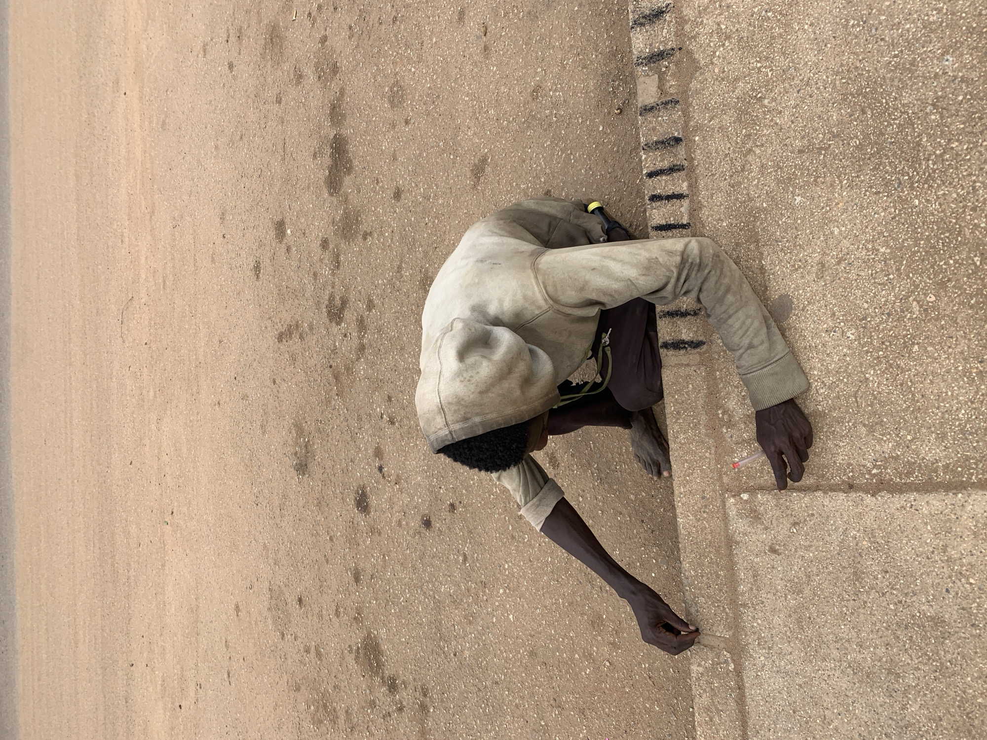 The image, rotated, captures an overhead view of a person crouched low to the ground next to a concrete curb.
The main subject is an individual, appearing to be male, with dark skin and short, dark curly hair. They are wearing a worn, light-colored hooded top that appears soiled, and dark trousers or shorts. A small yellow object is tucked into their clothing near the back of their neck.
The person is in a deep squat or crouch, intently focused on the ground and curb. In their right hand, they hold a small, clear, tube-like object with a red cap, which they seem to be using to make a mark or point at the horizontal surface of the concrete. Simultaneously, their left hand holds a smaller, light-colored object, possibly chalk, used to mark the side or top edge of the curb.
The setting is an outdoor space with a coarse, light brown ground surface, likely asphalt or gravelly concrete, showing some scattered darker stains. A light-colored concrete curb or low wall runs vertically along the right side of the frame. This curb features distinct short, black dashed lines painted intermittently on both its side and top surfaces. The natural lighting suggests it is daytime. No discernible text is visible.