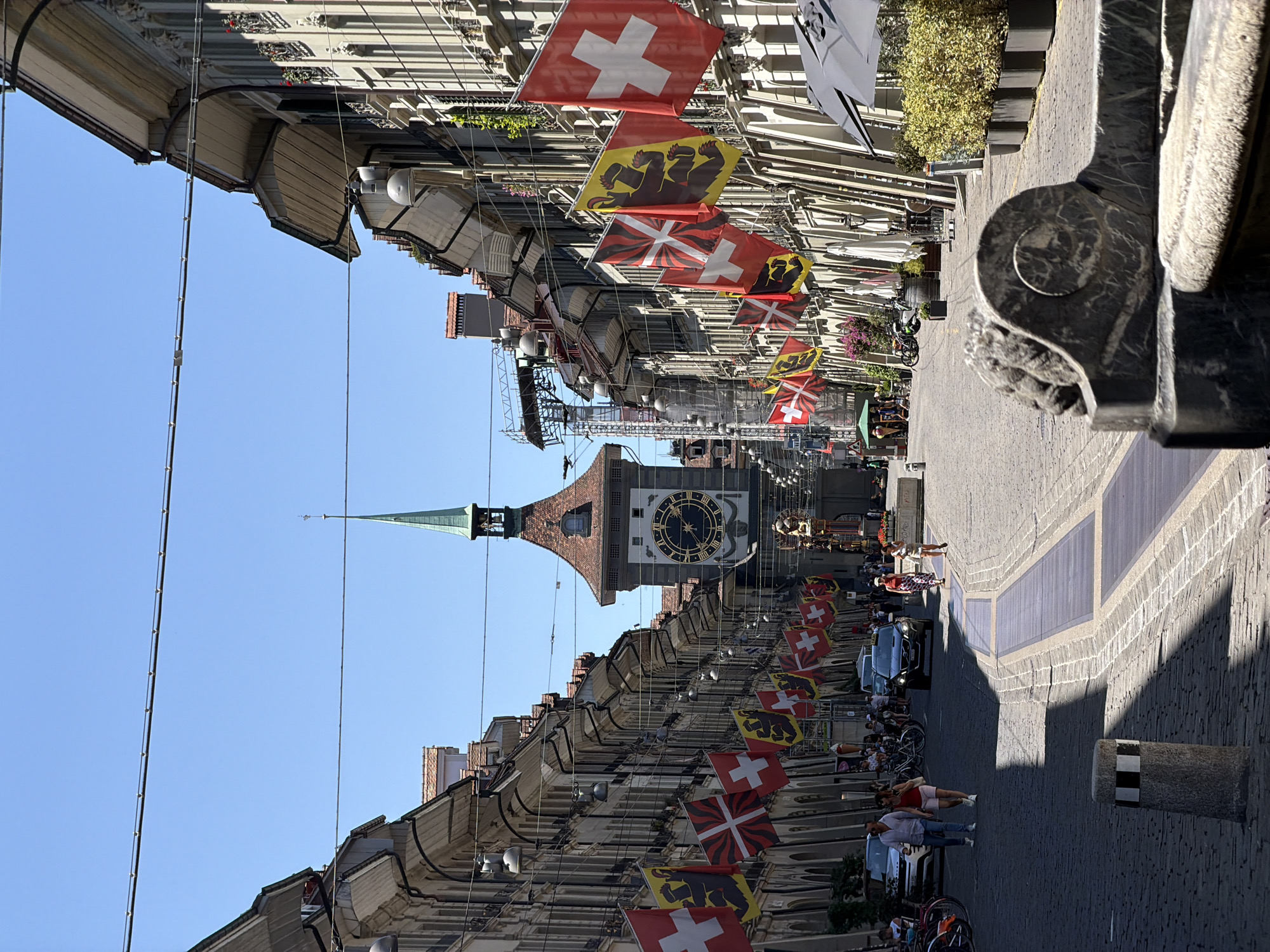 The image captures a street scene in Bern, Switzerland, likely in the Old Town, with the iconic Zytglogge clock tower dominating the background. The street is paved with cobblestones and lined with historic buildings featuring arched arcades and awnings. Numerous Swiss flags and cantonal flags (yellow with a black bear) are displayed along the street, suggesting a celebration or national holiday.
In the foreground, a stone fountain structure with a lion's head carving is visible. The street is moderately busy with people walking and some vehicles, including cars and bicycles. Several individuals are seen strolling along the sidewalks and crossing the street. A few parked cars are present, and a black SUV is driving down the street.
The Zytglogge tower, a medieval clock tower, stands prominently in the distance with its distinctive astronomical clock face. The sky is clear and blue, indicating a sunny day. The architecture of the buildings, the presence of Swiss flags, and the Zytglogge clock tower are strong indicators of the location being Bern, Switzerland.