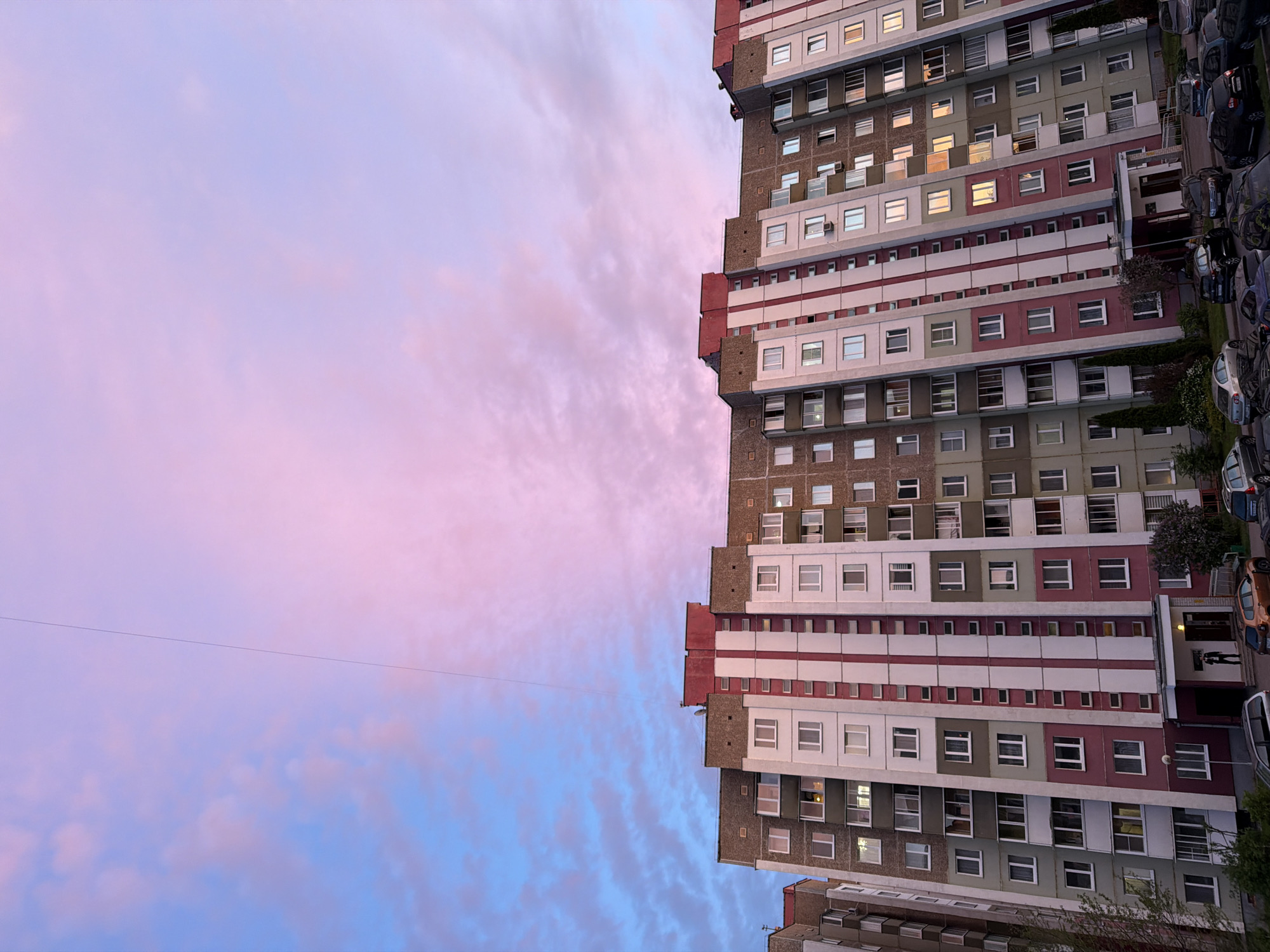 The image shows a tall apartment building in an unknown city. The building has a repeating pattern of white, grey, and burgundy stripes. Many windows are visible on the facade, some reflecting sunlight. A number of cars are parked along the side of the building. Above the building is a sky with a blend of pink, purple, and blue hues, with streaky clouds. An electrical wire is stretching across the top side of the image.