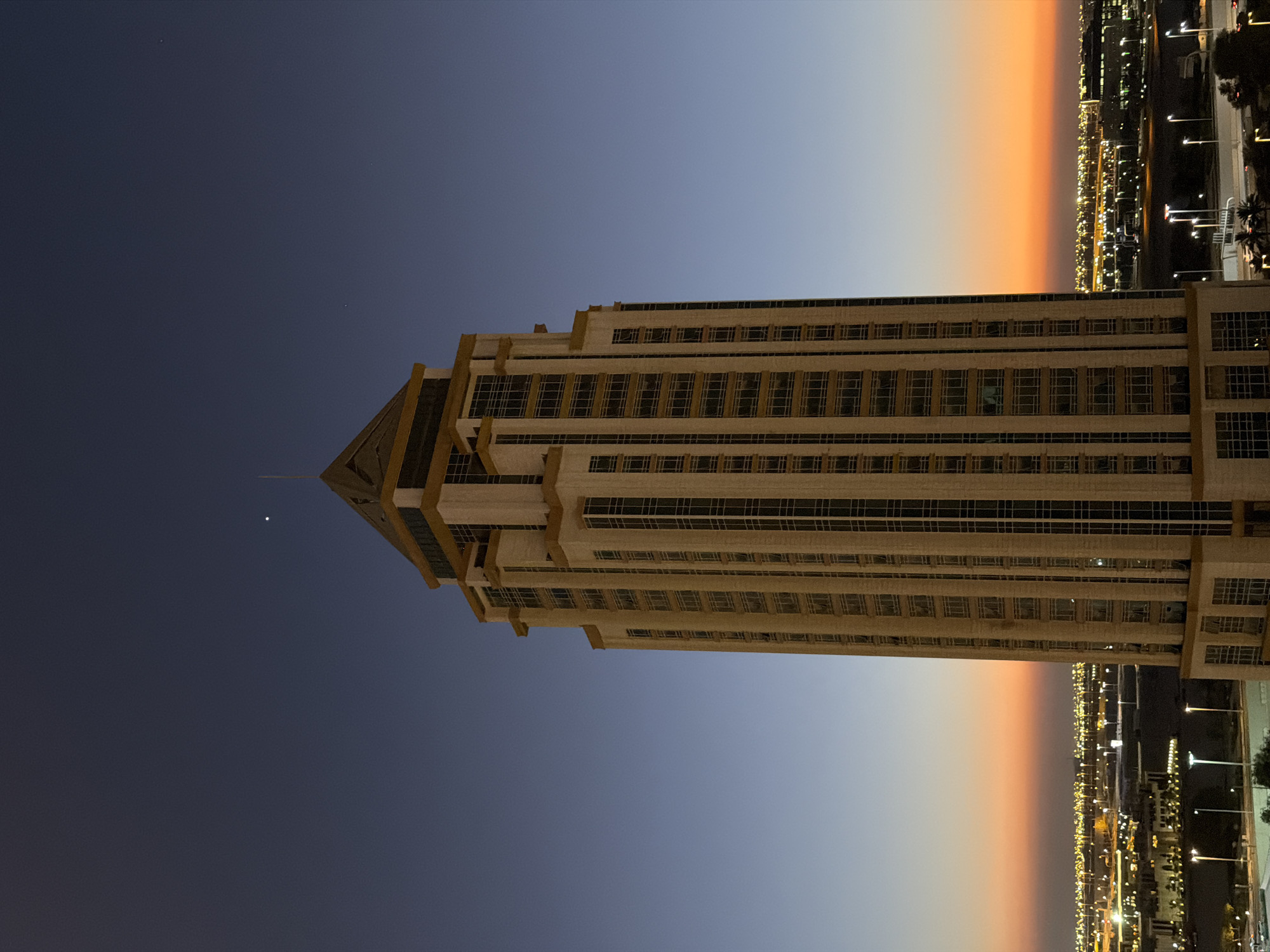 This is a vertical, eye-level, full-shot photograph taken at dusk, capturing a tall, multi-story skyscraper in the foreground. The skyscraper, predominantly beige with dark windows, rises majestically against a gradient sky that transitions from a deep indigo at the top to a warm orange and yellow hue near the horizon. A single bright star is visible in the upper portion of the sky. The skyscraper has a stepped architectural design, with setbacks on higher floors and a distinctive pointed roof topped with a thin spire. Below the skyscraper, a sprawling cityscape is illuminated by numerous lights, suggesting an urban environment. Roads with streetlights are visible, indicating infrastructure. The overall scene depicts a modern city at twilight.